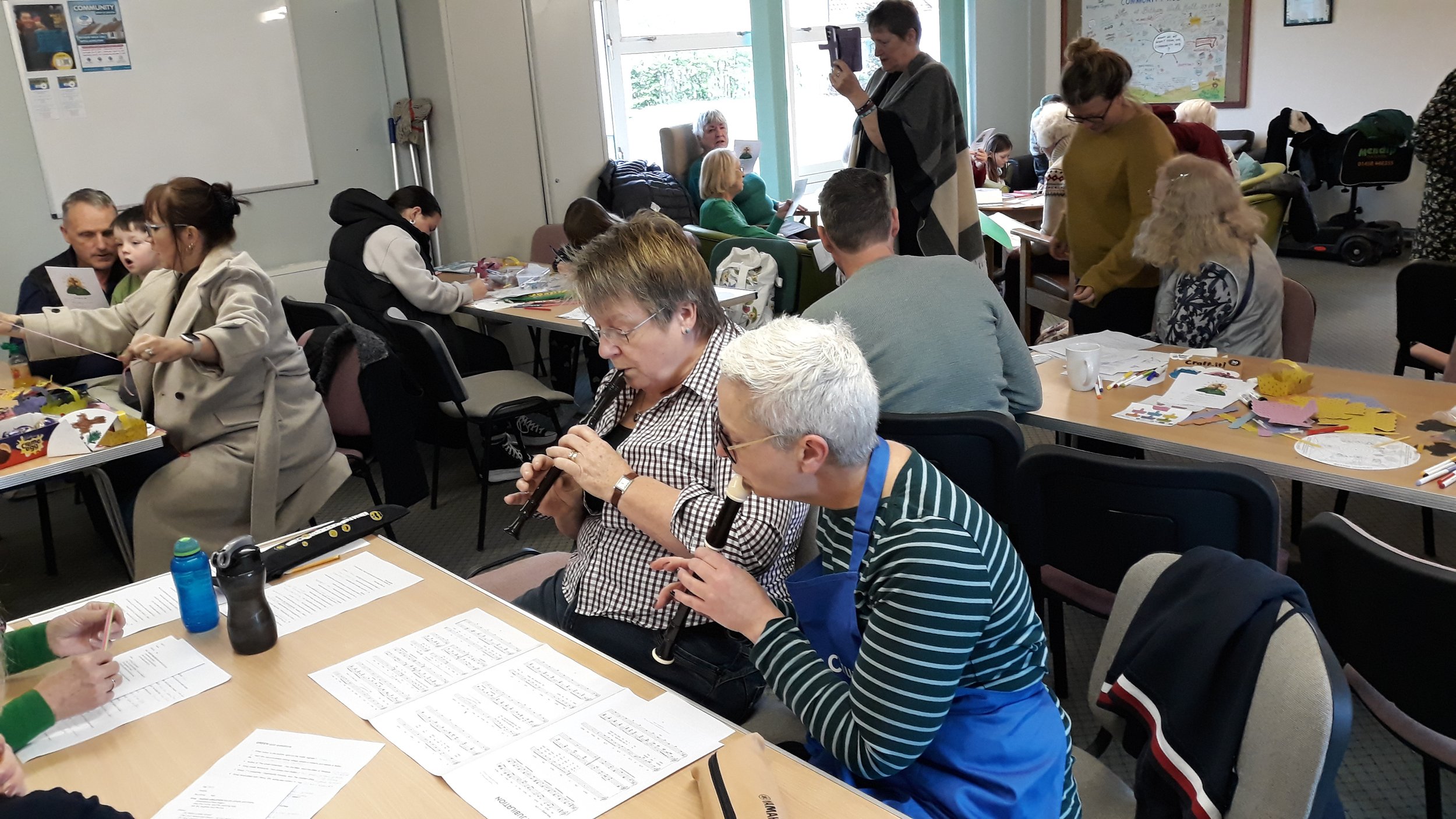 People in a classroom or community center participating in various activities, including children and adults sitting at tables working on crafts, some playing musical instruments, and others engaged in conversations or taking photos.