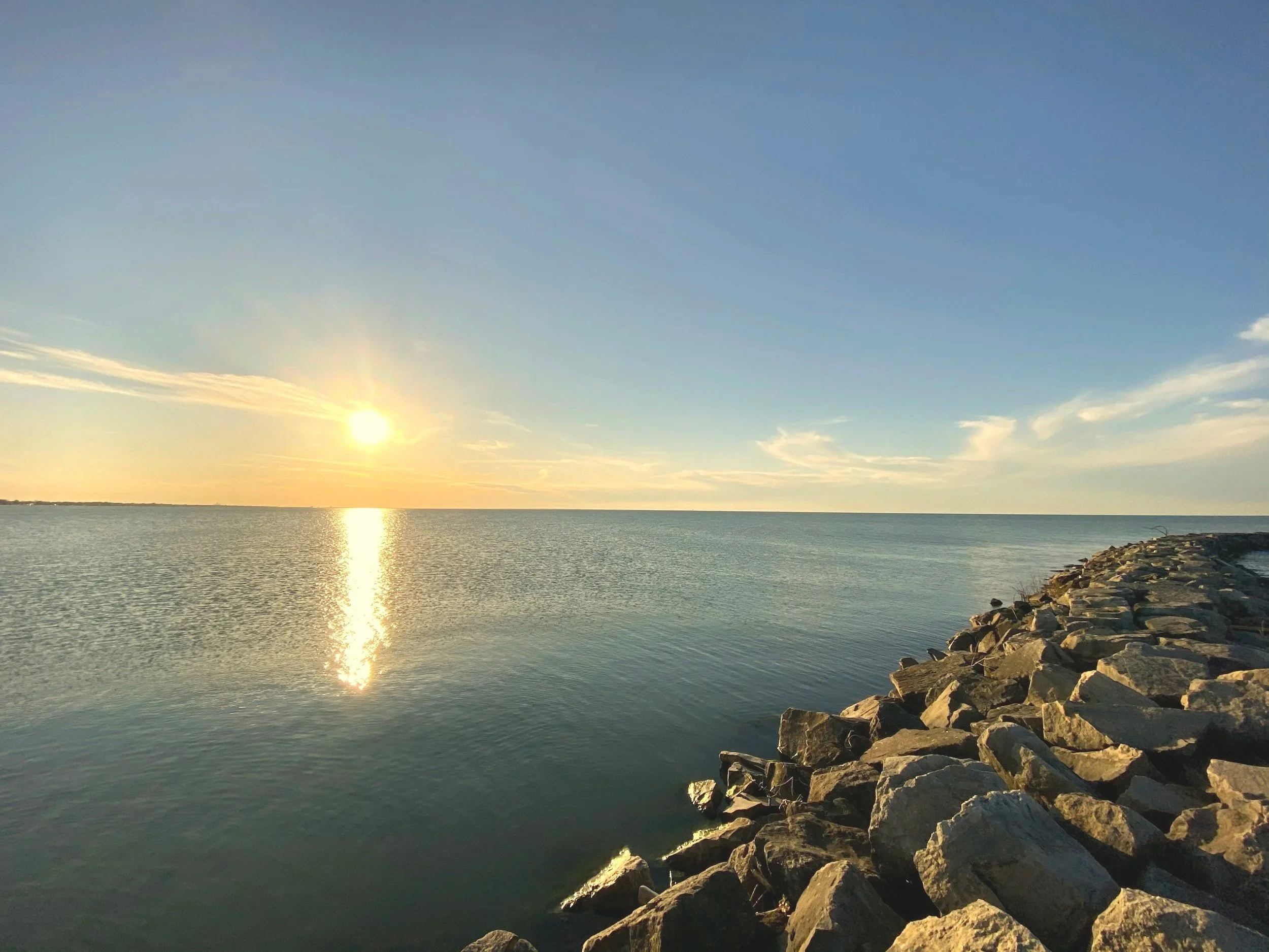 A scenic view of the sunset over calm water, with rocks lining the shoreline on the right, and a clear sky with few clouds.