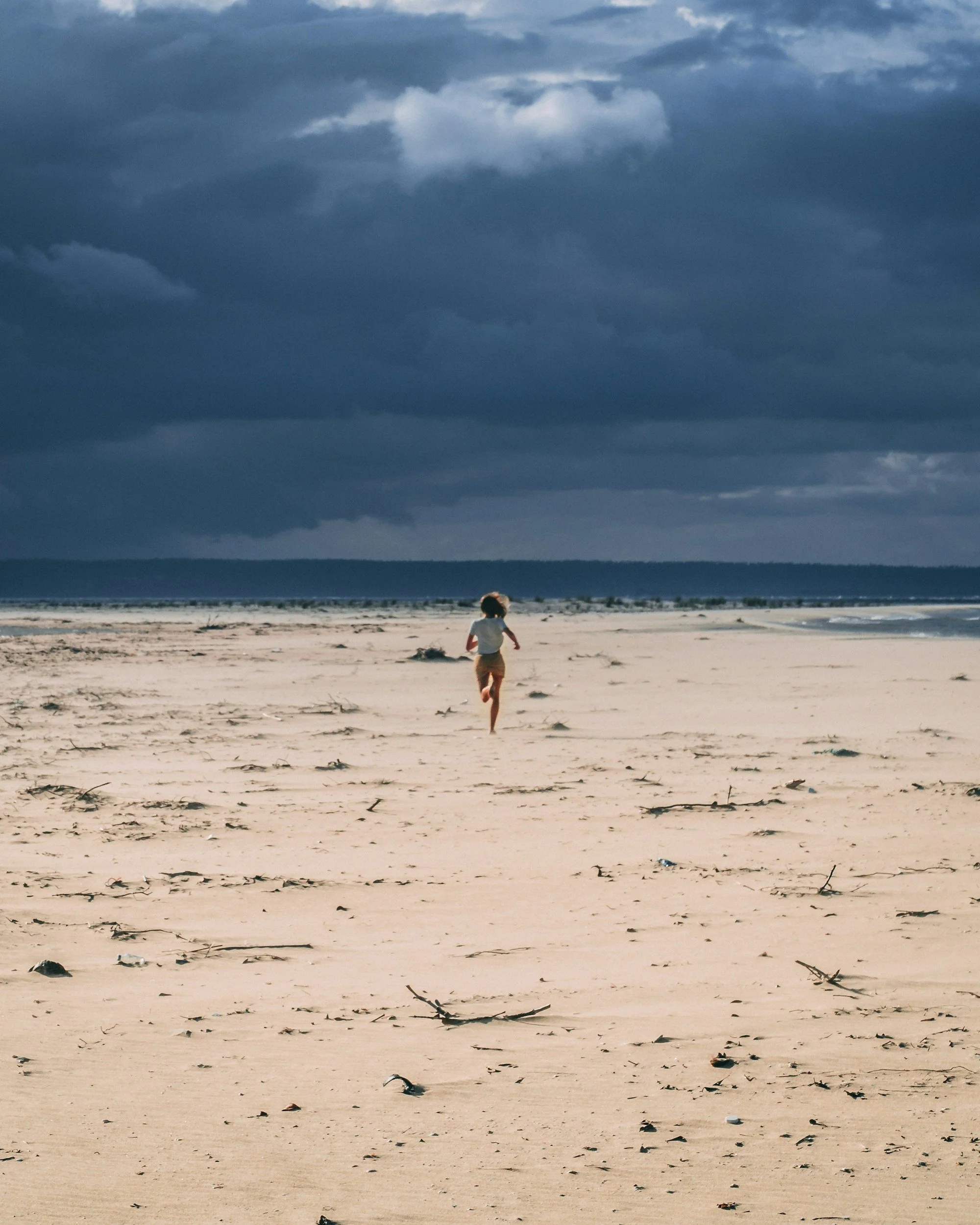 A child running on a sandy beach under a dark, cloudy sky.