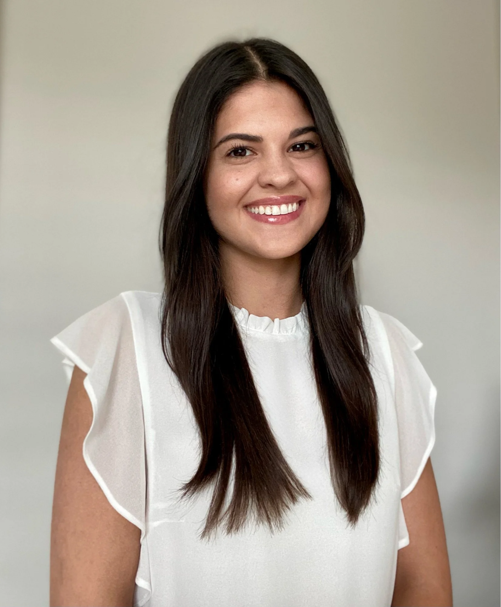 Portrait of a woman with long dark hair, smiling, wearing a white blouse, against a plain background.