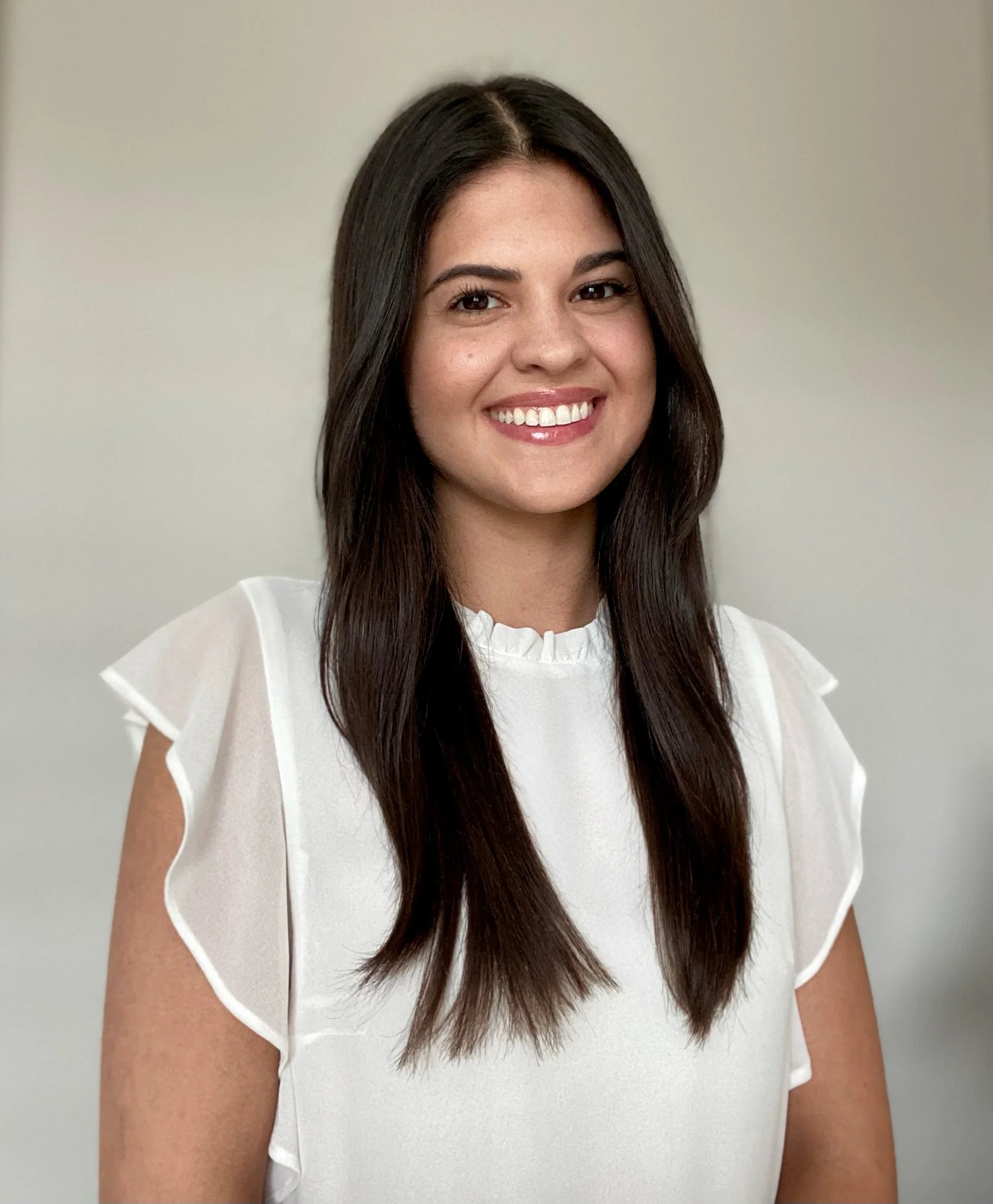 A woman with long dark brown hair smiling, wearing a white blouse with ruffled sleeves, standing against a plain light-colored background.