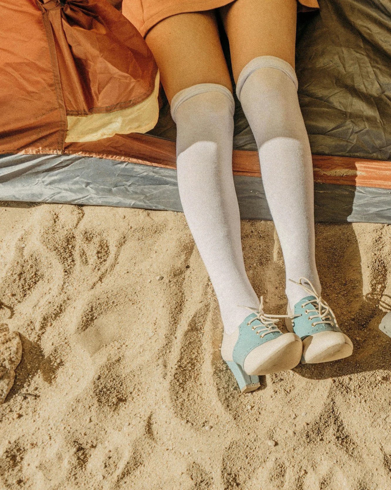 Person lying on a bed with legs extended. They are wearing white knee-high socks and light blue shoes with white soles and laces. The bed has brown and gray bedding, and the floor is sandy.