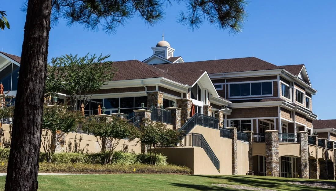 Large multi-story amenity center, multiple balconies with railing, and a large sloped roof with a small cupola, surrounded by greenery and trees under a clear blue sky.