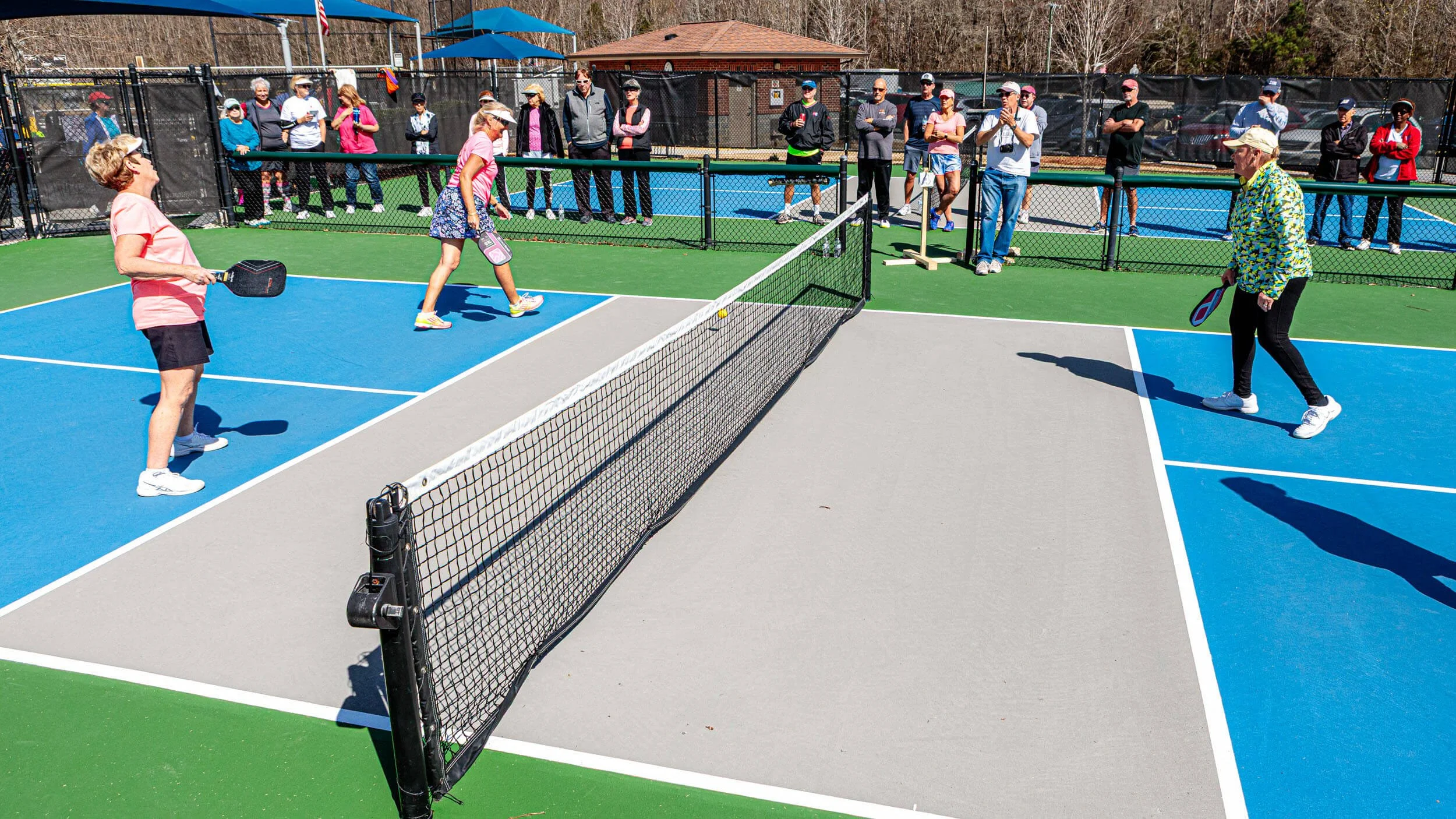 People playing pickleball on a colorful outdoor court with a net, surrounded by spectators watching.