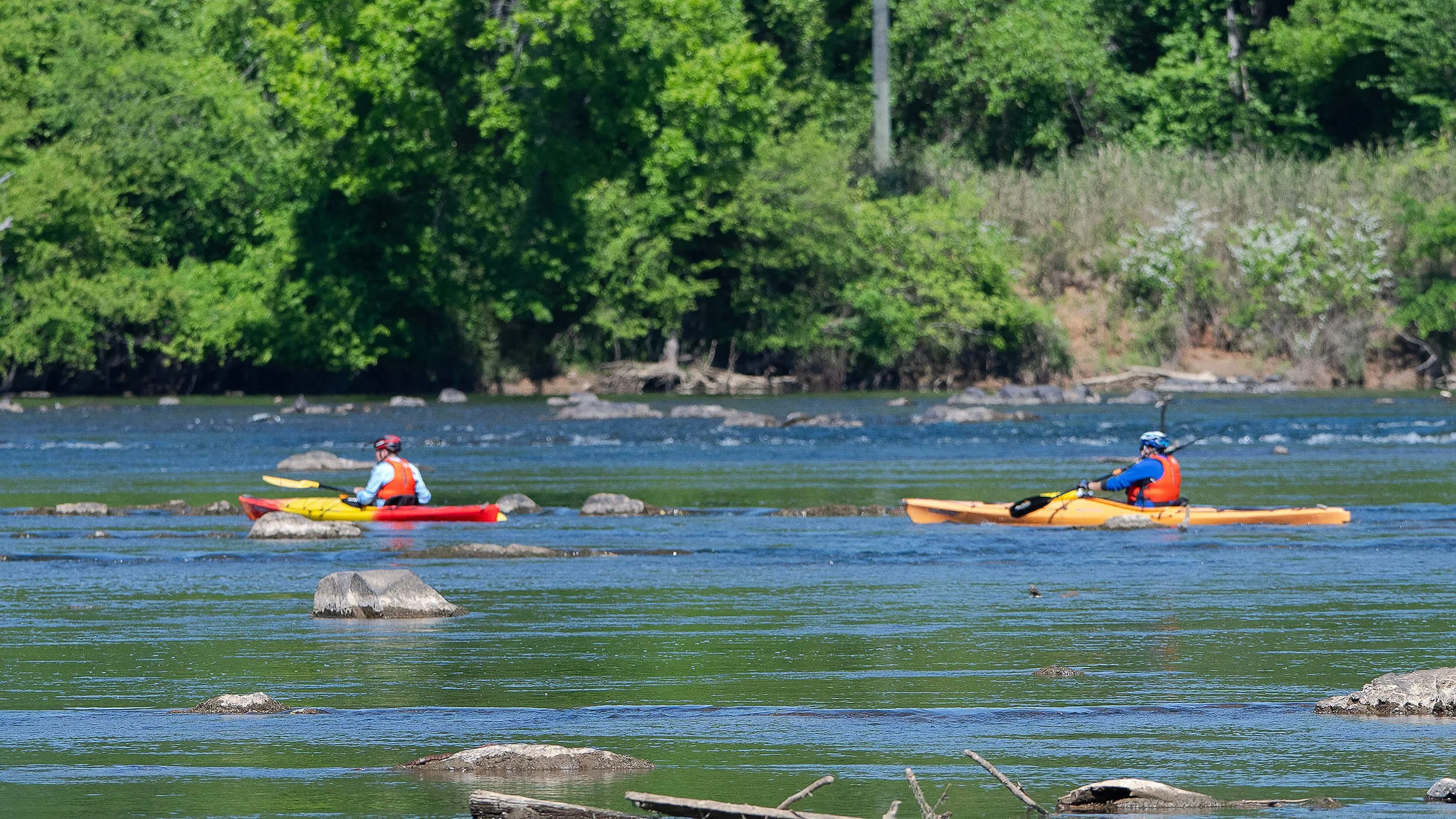 Two people kayaking on the Catawba  river with green trees in the background, rocky riverbed, and clear weather.