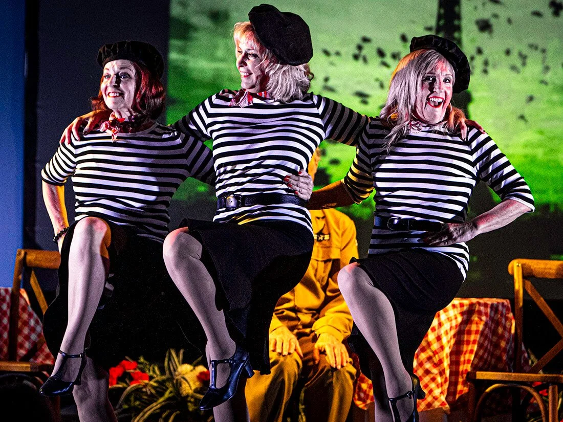 Three women dressed as mime performers in black and white striped shirts, black skirts, and berets performing a dance on stage with a green background and an audience member dressed in yellow seated behind them.