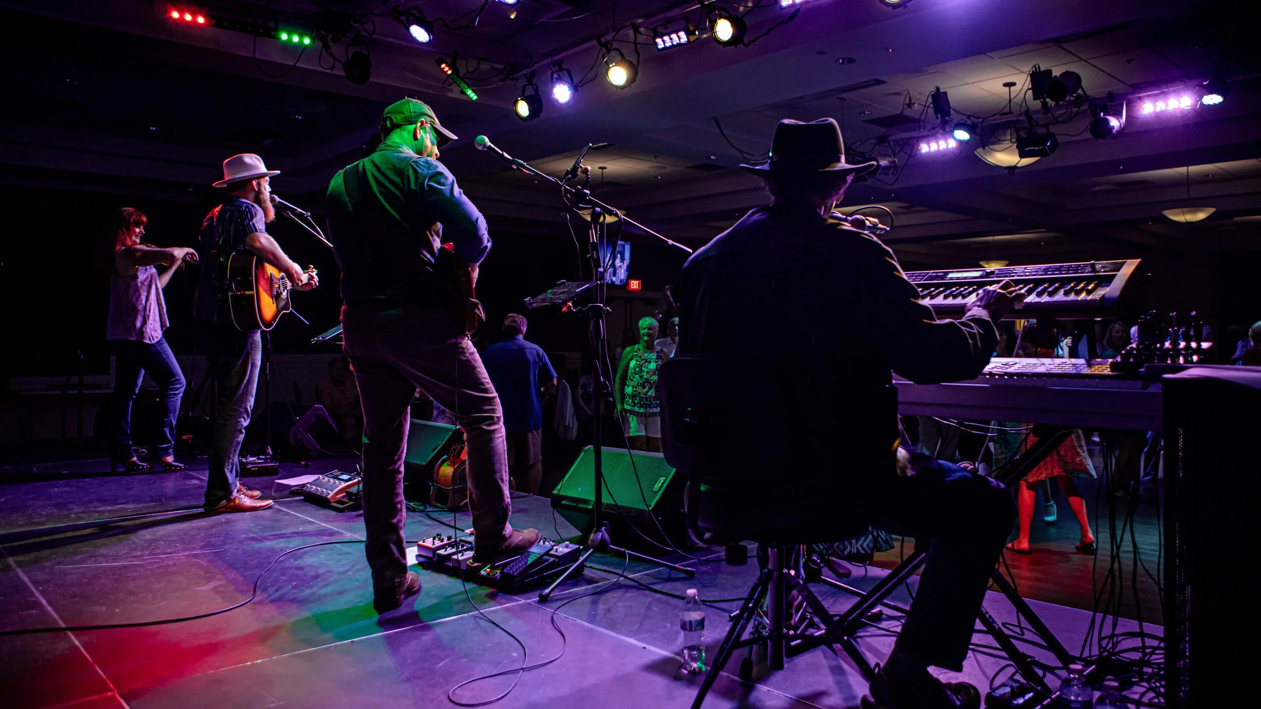 A band performing on stage with colorful lighting, including musicians playing guitars and a keyboard, and audience members dancing and enjoying the music.
