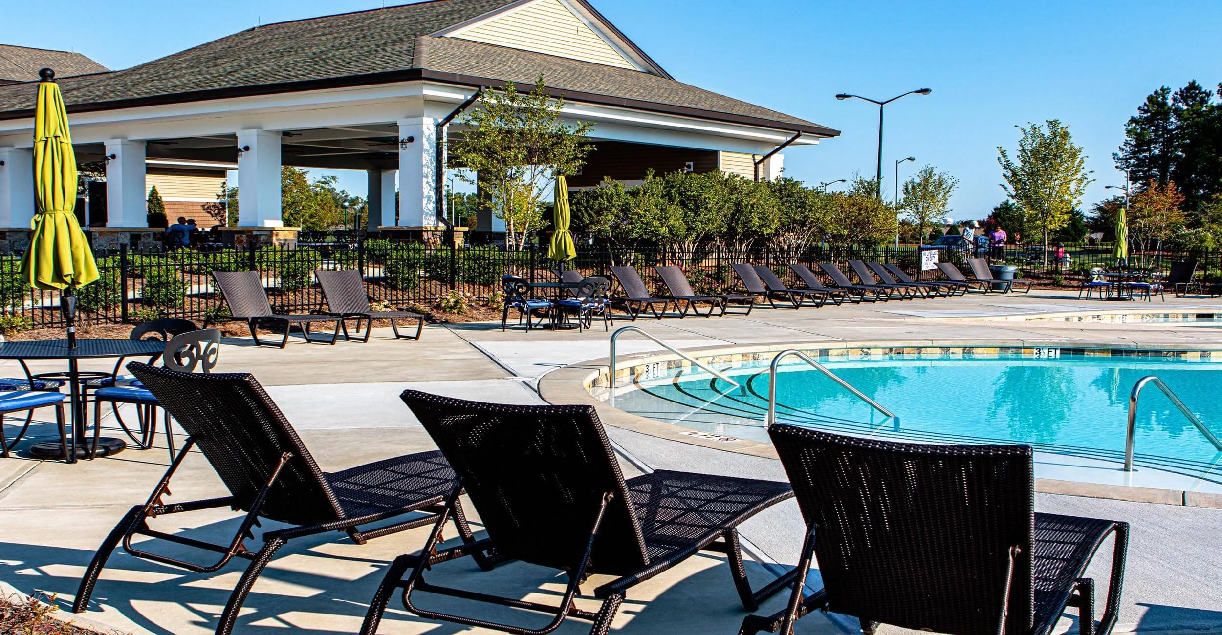 An outdoor swimming pool area with lounge chairs, tables, and umbrellas, surrounded by a black fence with trees and a building in the background.