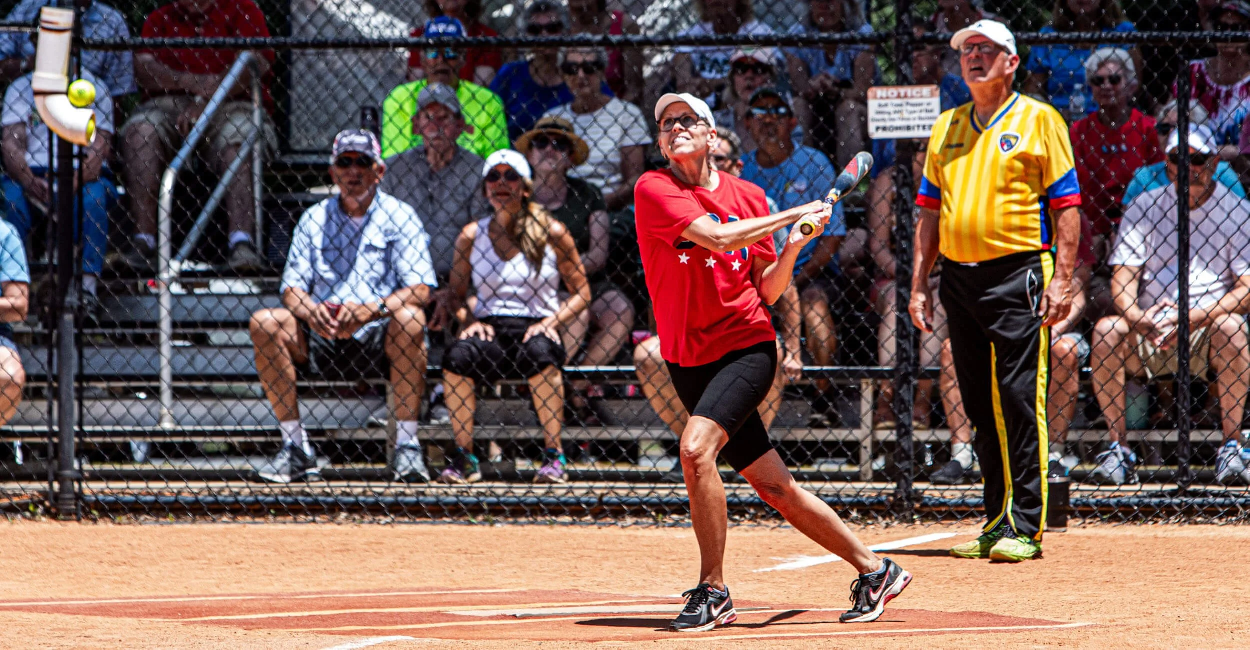 Active adults in Sun City playing softball, and a crowd of spectators behind a chain-link fence watching the game.
