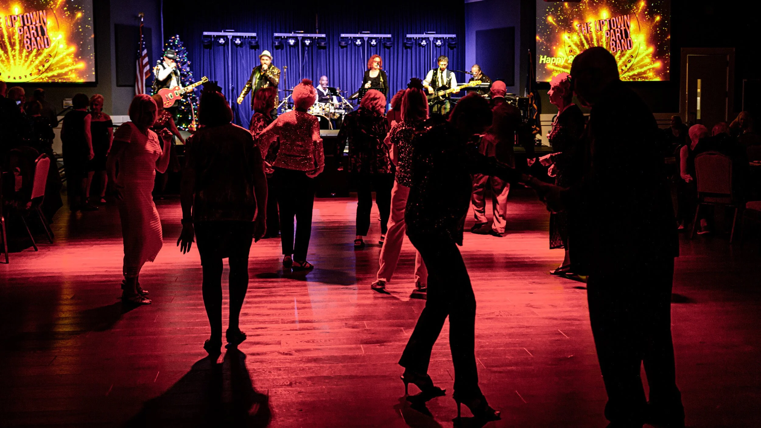 People dancing on a wooden floor in front of a live band on stage with a curtain backdrop, and two screens displaying colorful graphics and text.