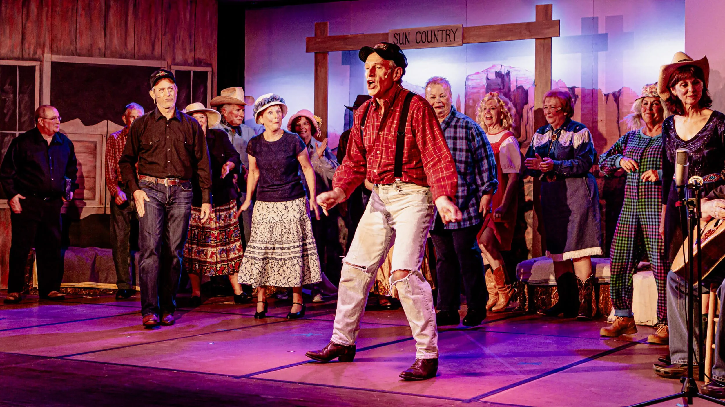A group of performers dressed in country-themed costumes on stage, with one man in the center singing or speaking, under a sign that reads 'Sun Country', with wooden set pieces and a painted backdrop of a desert landscape.