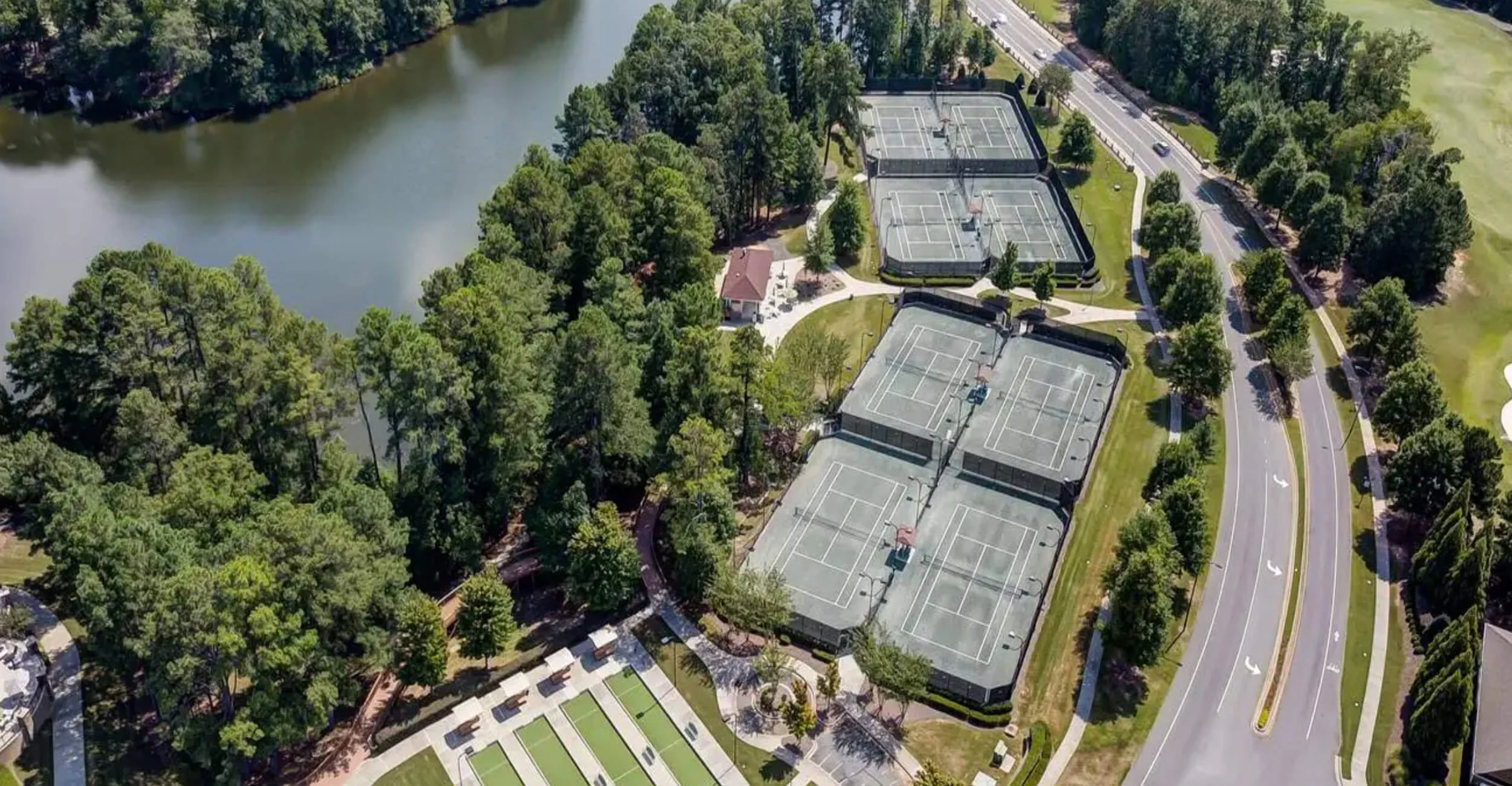 Aerial view of Carolina Lakes Har-tru tennis courts, with a lake, surrounding trees, a small building, parking lot, and roads.