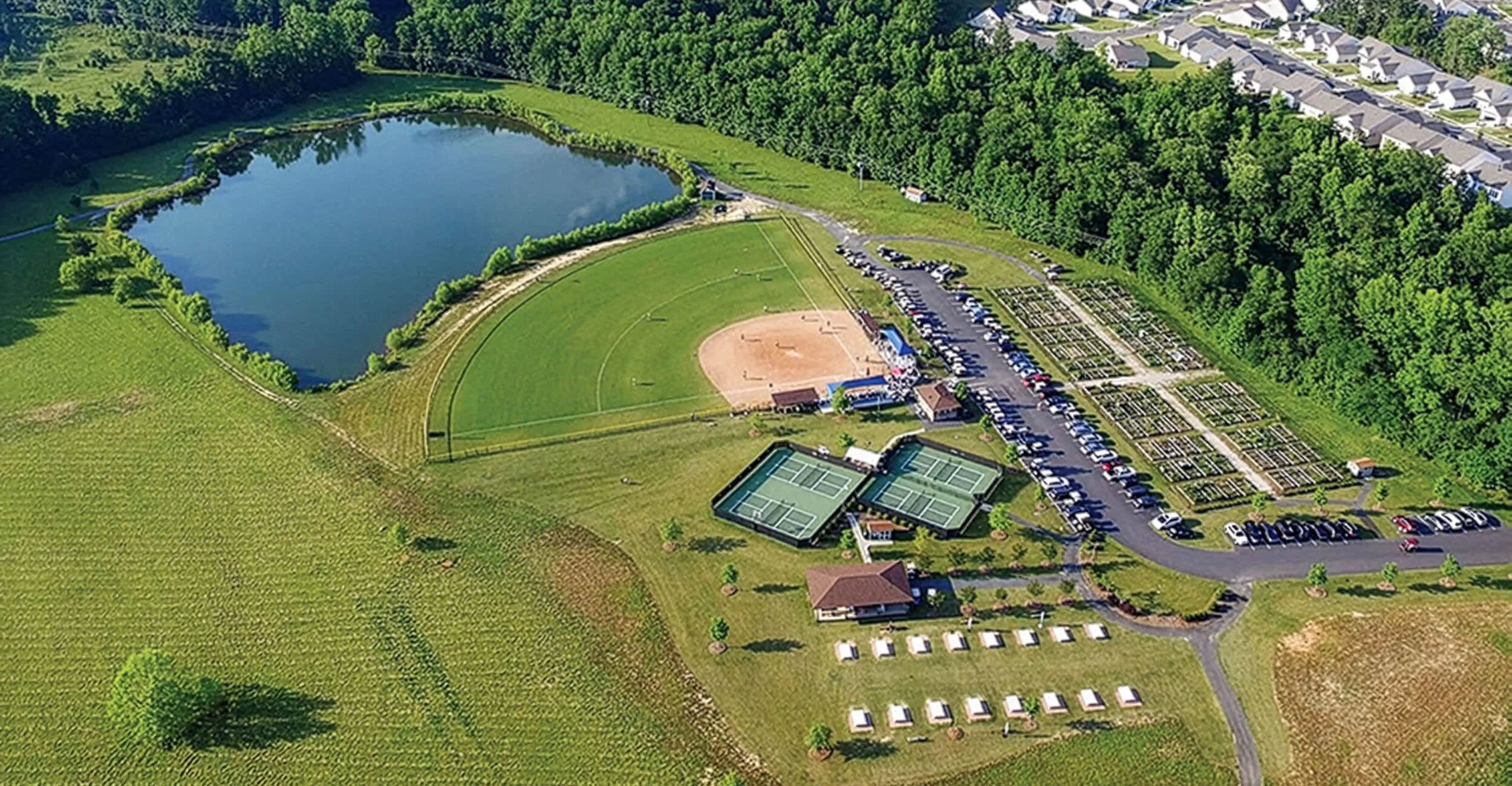 Aerial view of Waters Edge park, and residential area featuring a pond, pickleball courts, baseball field, parking lot, and green spaces surrounded by trees.