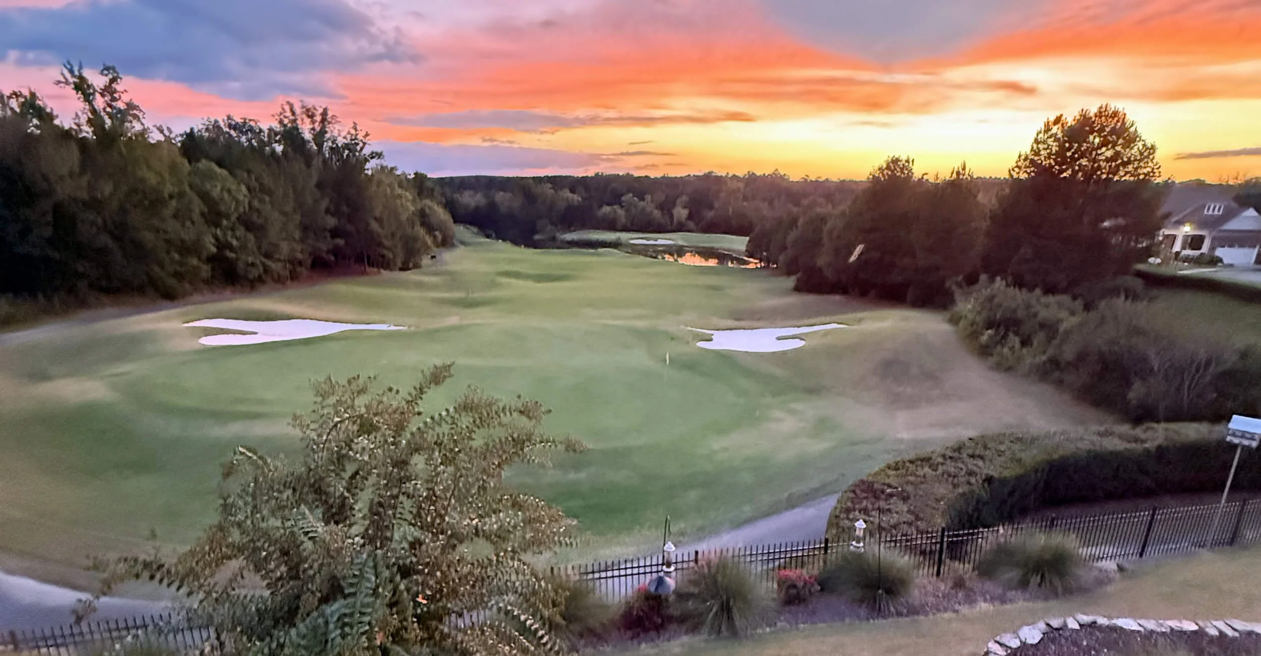 Carolina Lakes golf course at sunset with a colorful sky, sand traps, water hazards, and surrounding trees and houses.