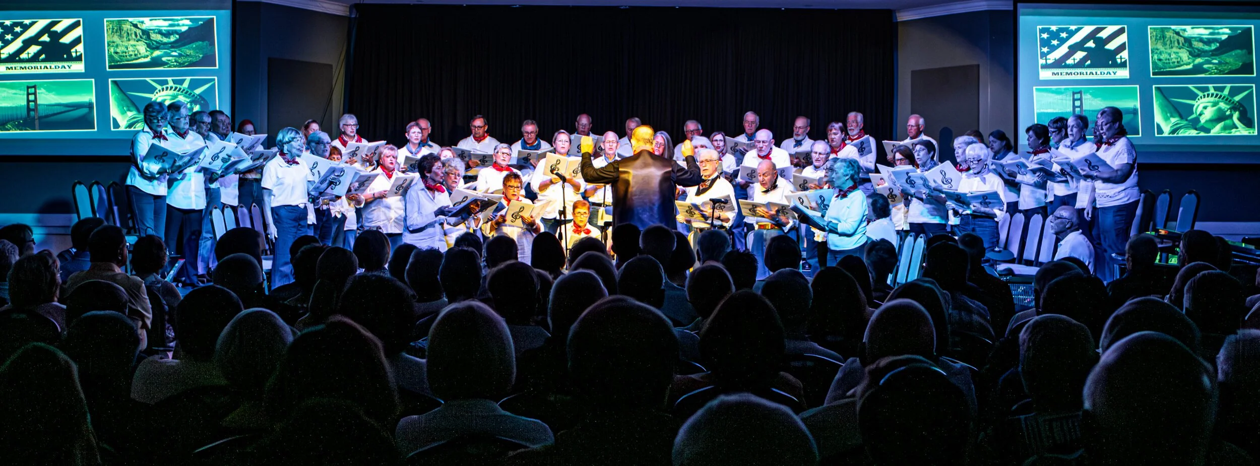 Choir performing on stage with a conductor, in front of an audience, with patriotic images of American landmarks and flags projected on screens. This is from The Lodge ballroom.