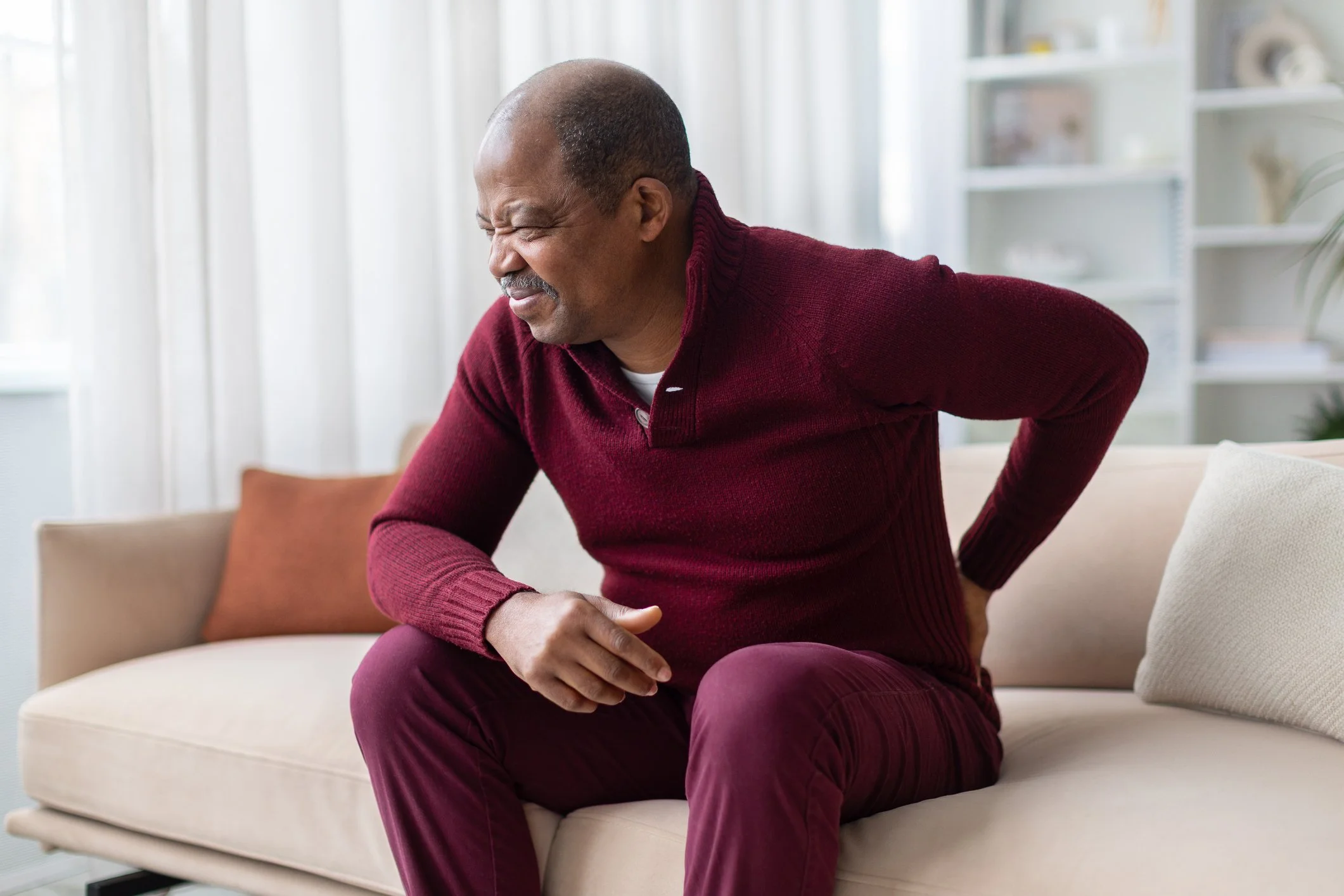 Man sitting on a beige sofa holding his lower back with a pained expression, wearing a red sweater and pants in a living room. Aortic Disease. Complex Aortic Disease. Venous Pathologies. Mesenteric  Ischaemia.