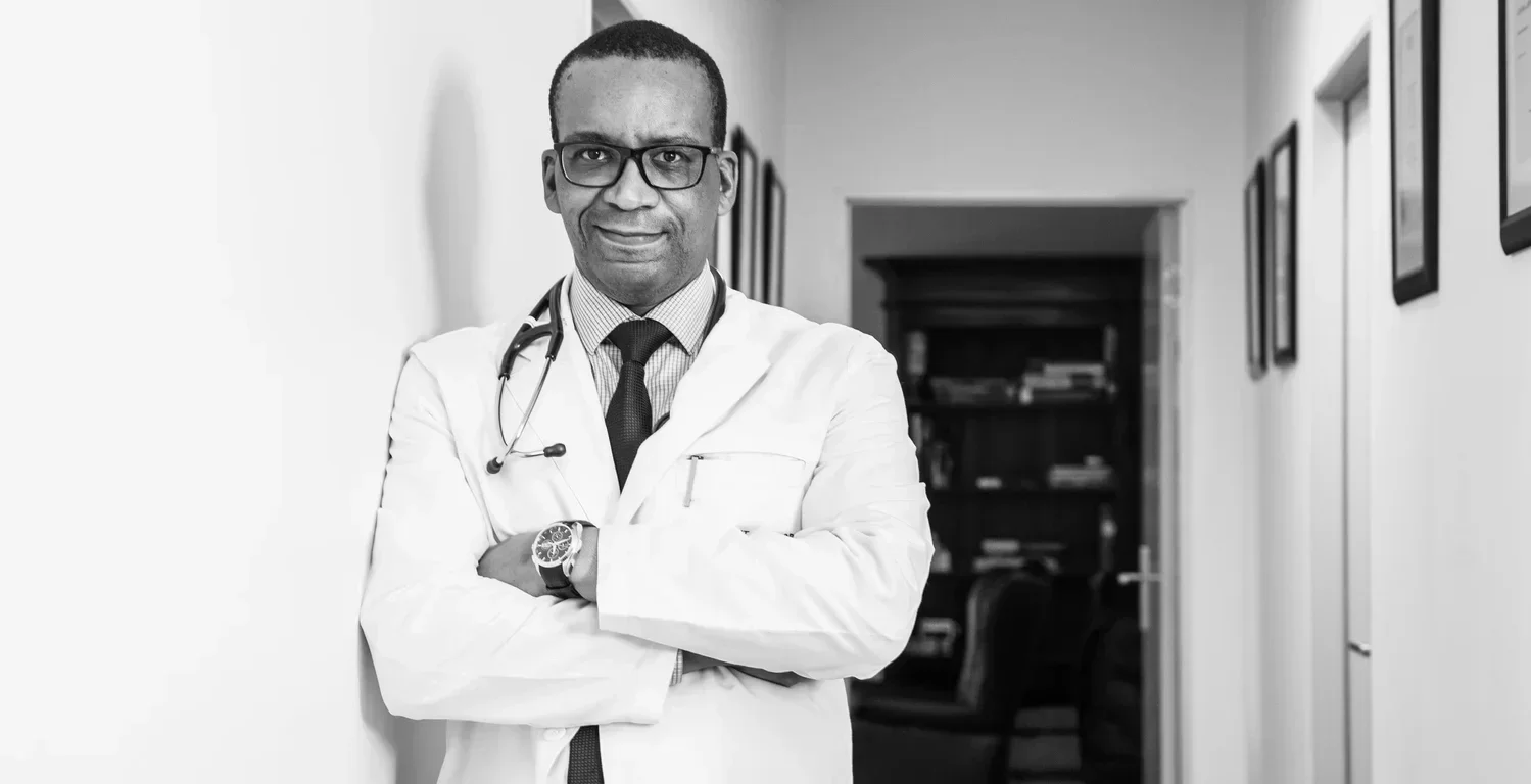 A male doctor with glasses and a stethoscope around his neck, wearing a white coat, in a professional setting.