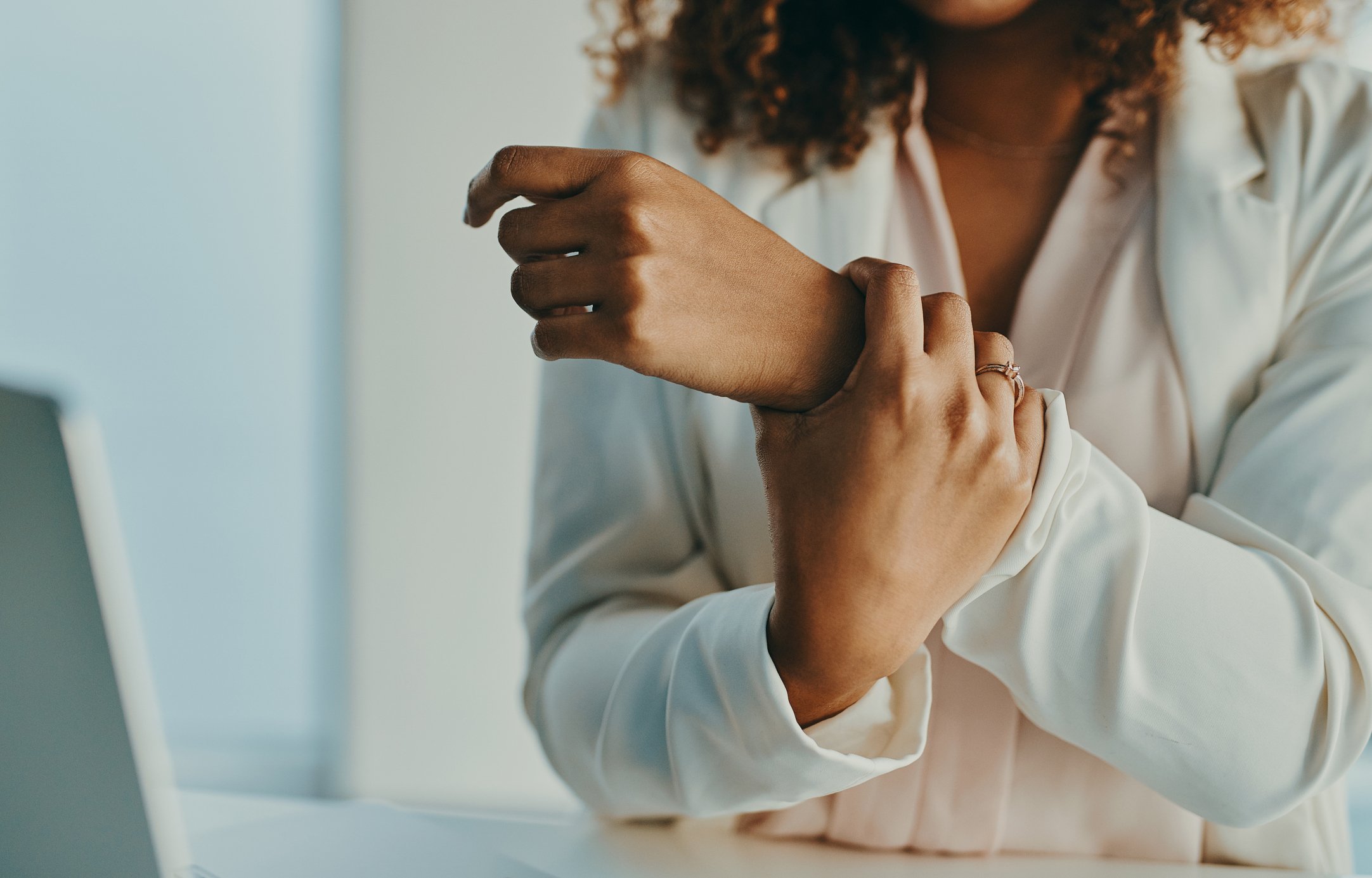 A woman with curly hair wearing a white blouse touching her wrist with her other hand, sitting at a desk.