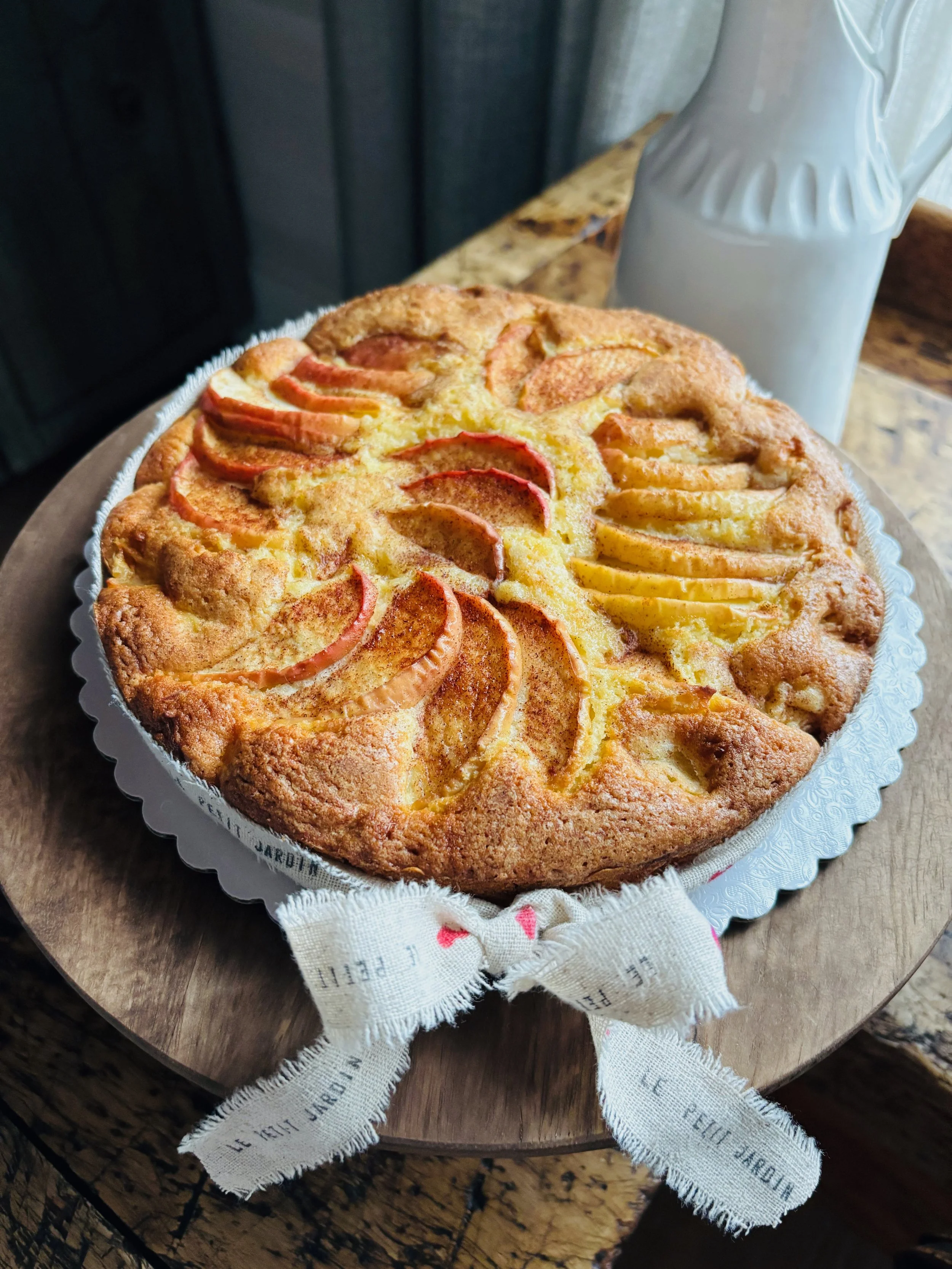 A freshly baked fruit tart with sliced peaches and apples on top, placed on a wooden round table with a white ceramic pitcher beside it.