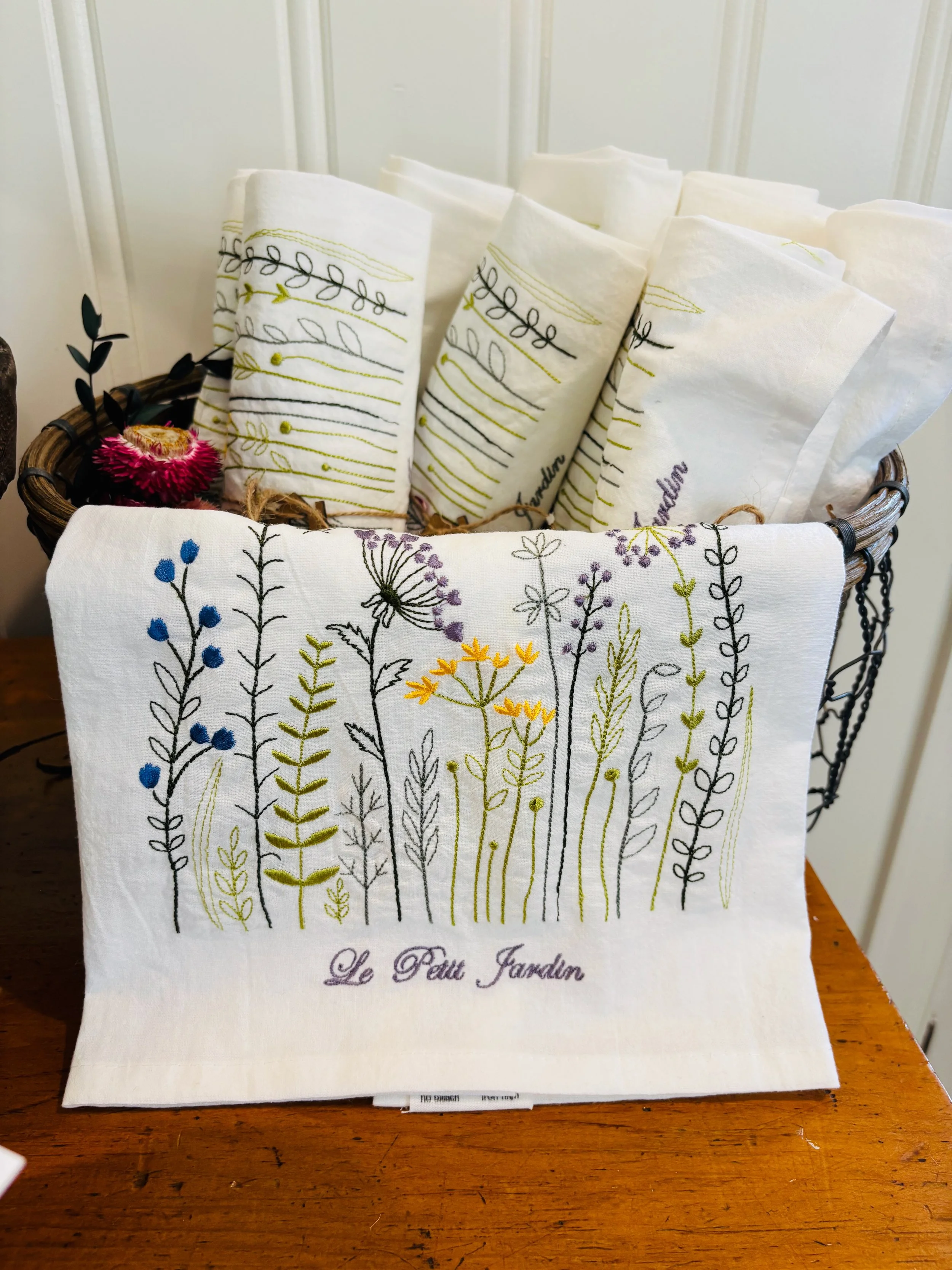 A basket with multiple white cloth napkins embroidered with colorful floral and leaf patterns, with the words 'Le Petit Jardin' embroidered on one napkin, placed on a wooden surface.
