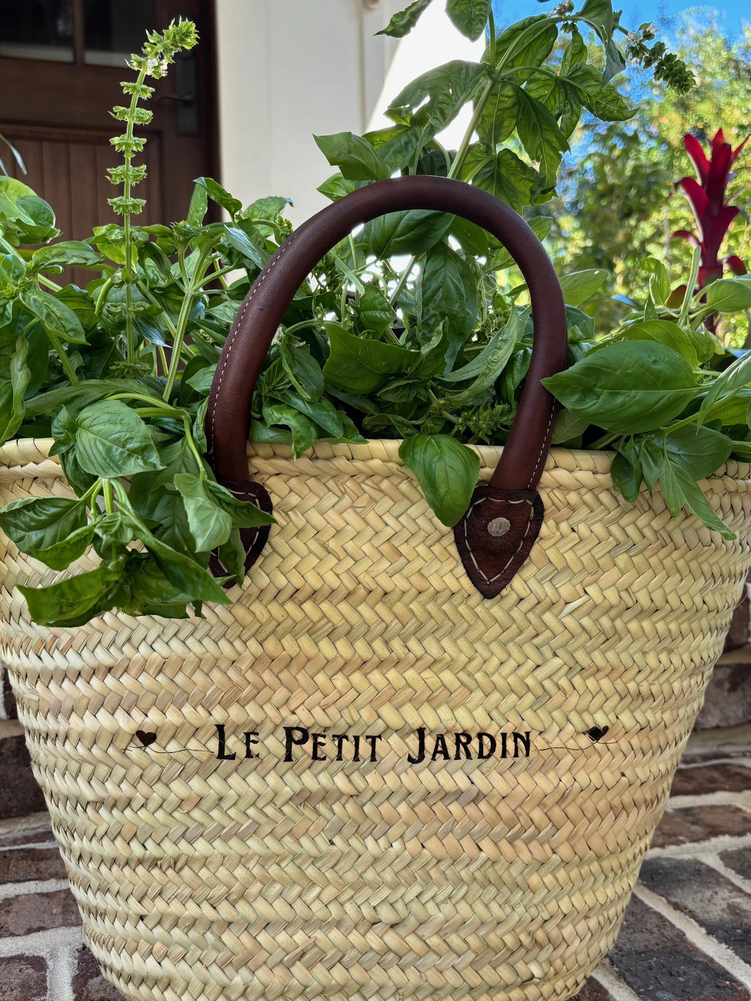 A woven basket with leather handles and the words "Le Petit Jardin" written on it, filled with green plants and flowers.