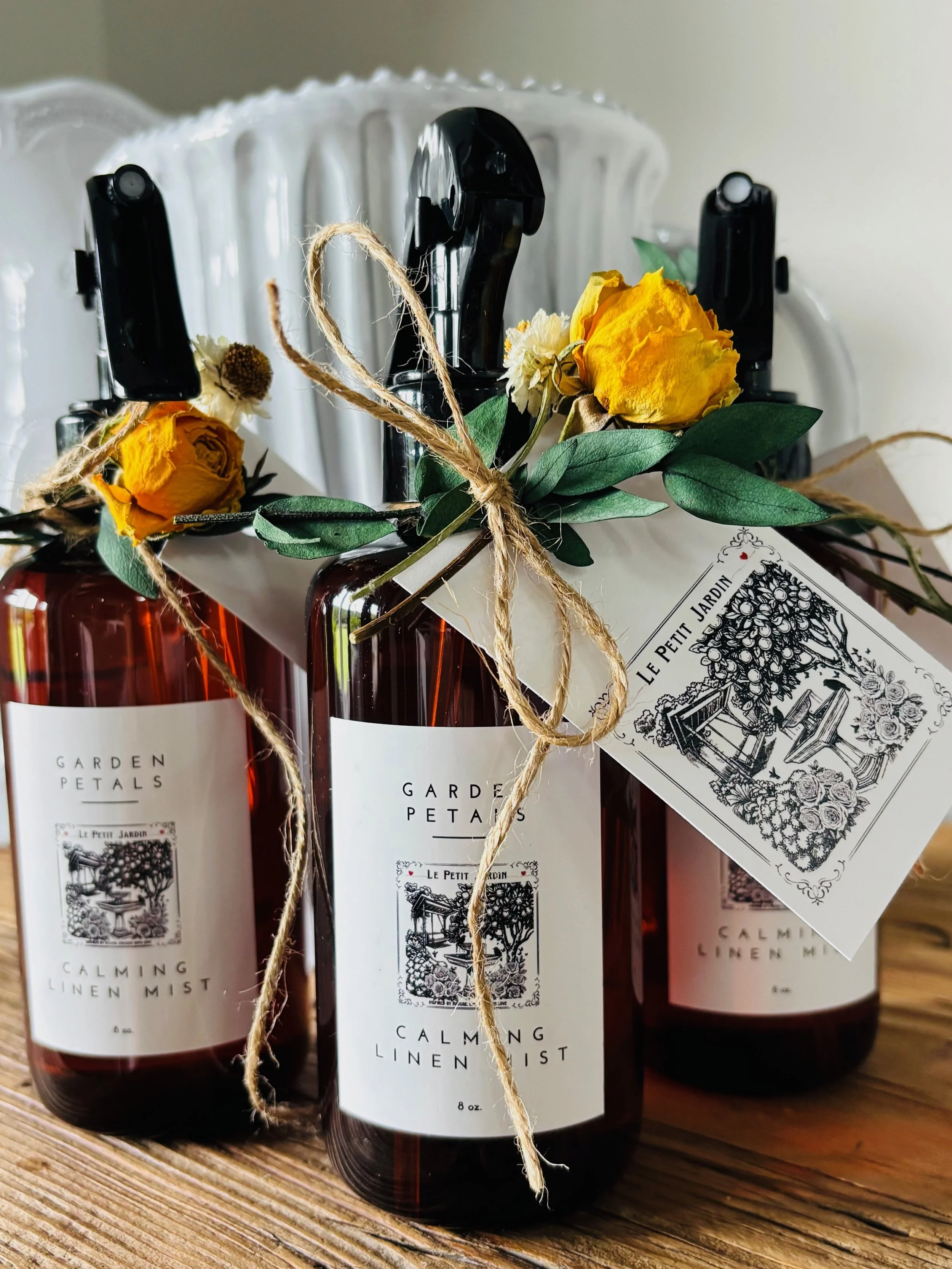 Three amber glass spray bottles with white labels reading 'Garden Petals Calming Linen Mist,' decorated with yellow and white dried flowers and green leaves tied with twine; placed on a wooden surface with a white ceramic object in the background.