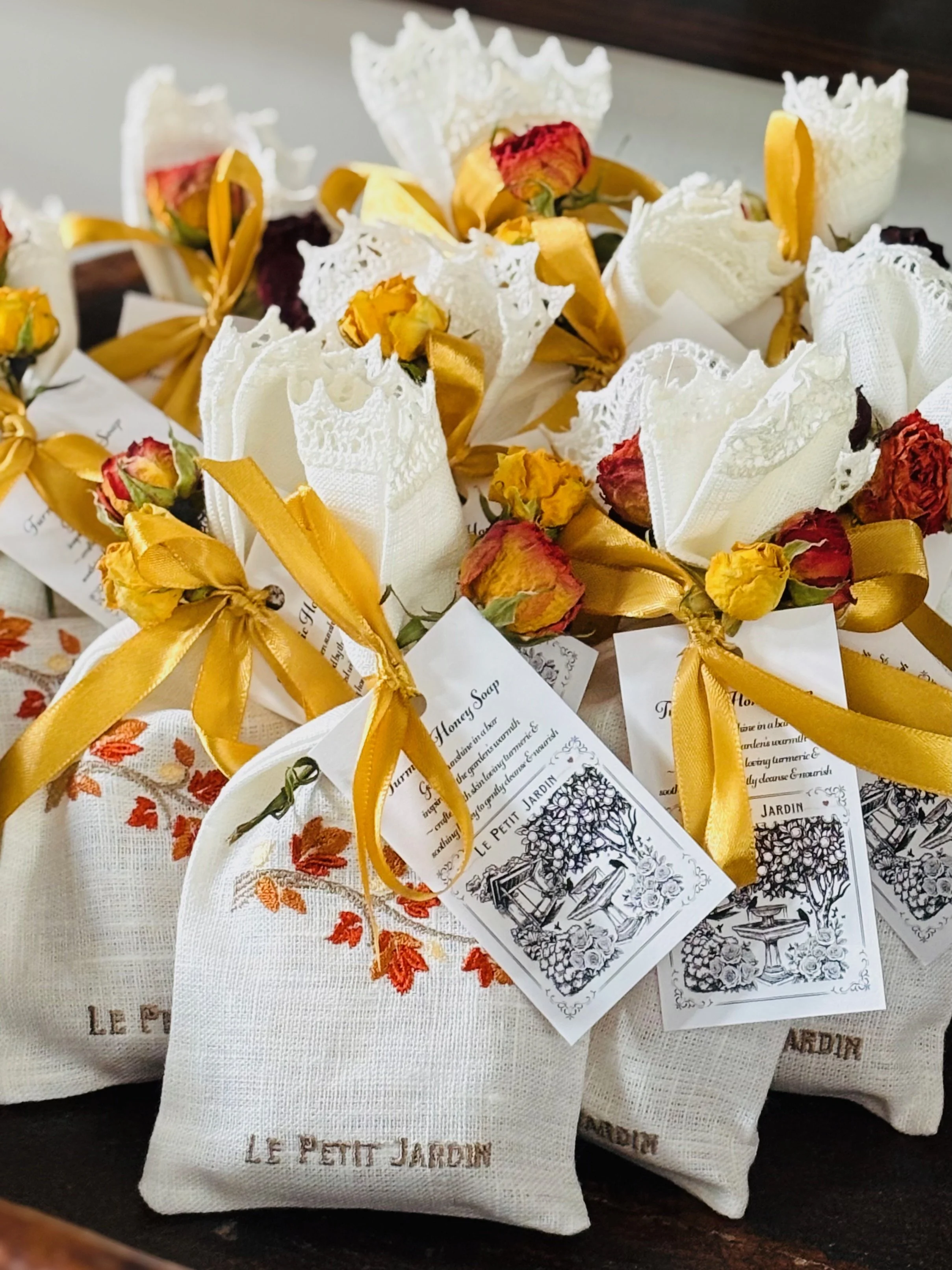 Decorative flower bouquets with dried roses tied with yellow ribbons, placed inside white cloth pouches embroidered with orange floral designs that read 'Le Petit Jardin'.