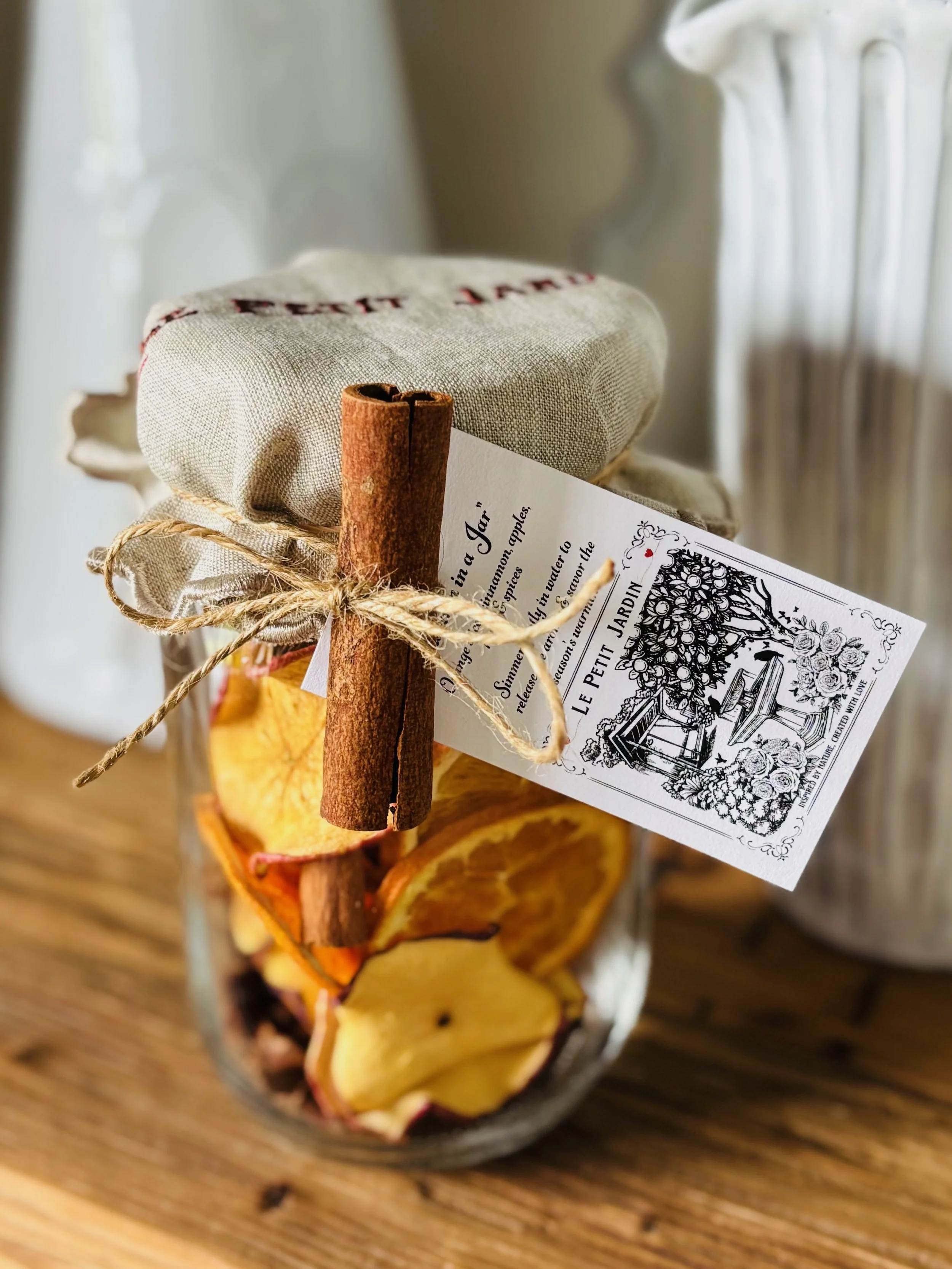 A glass jar filled with dried apple slices, topped with a fabric cover secured by a twine bow, with a cinnamon stick and a decorative tag attached.