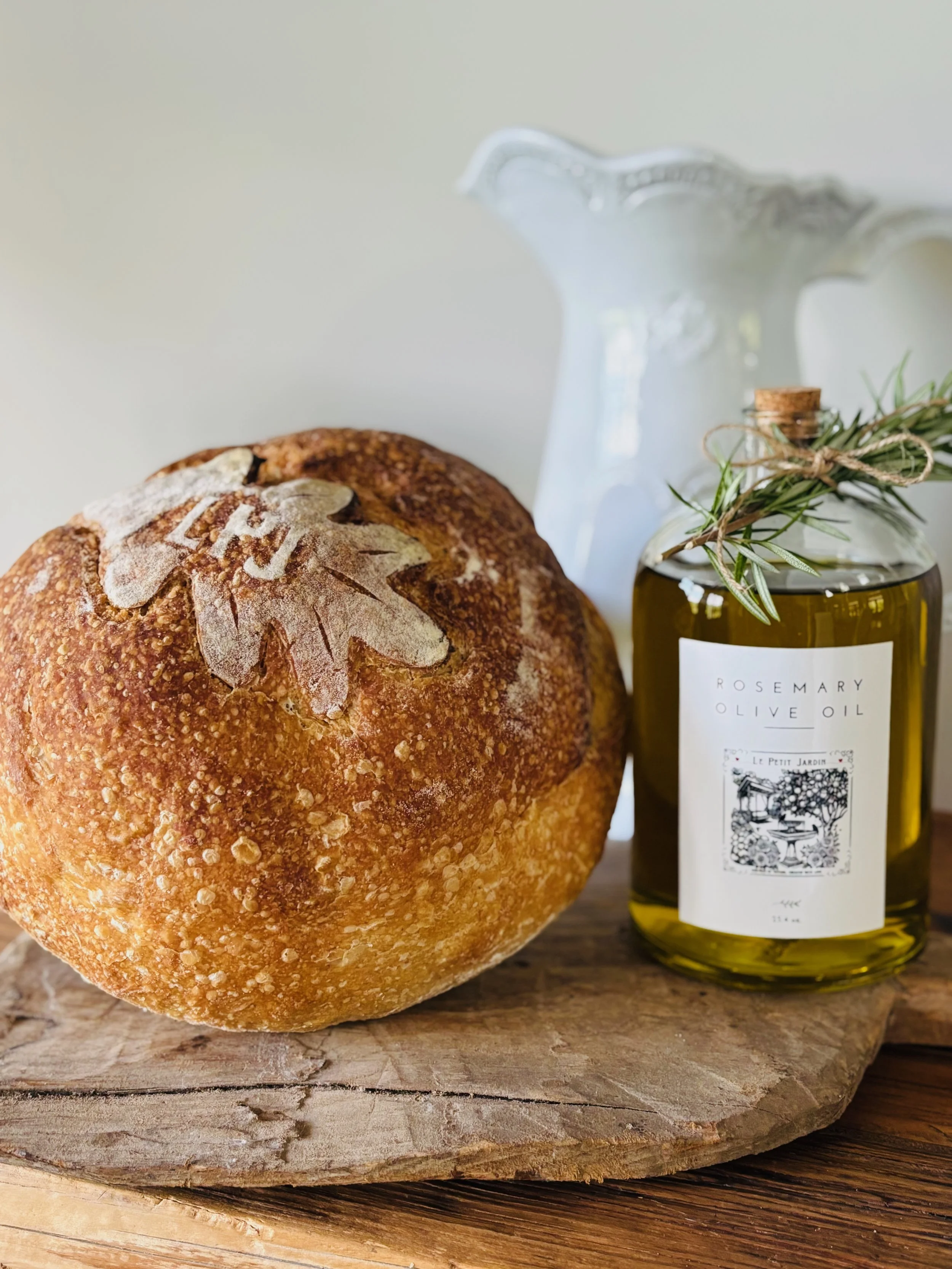 A loaf of rustic bread with a leaf-shaped decoration and the word 'LÉG' on top, placed on a wooden surface next to a bottle of rosemary olive oil with a sprig of rosemary, against a blurred light background.