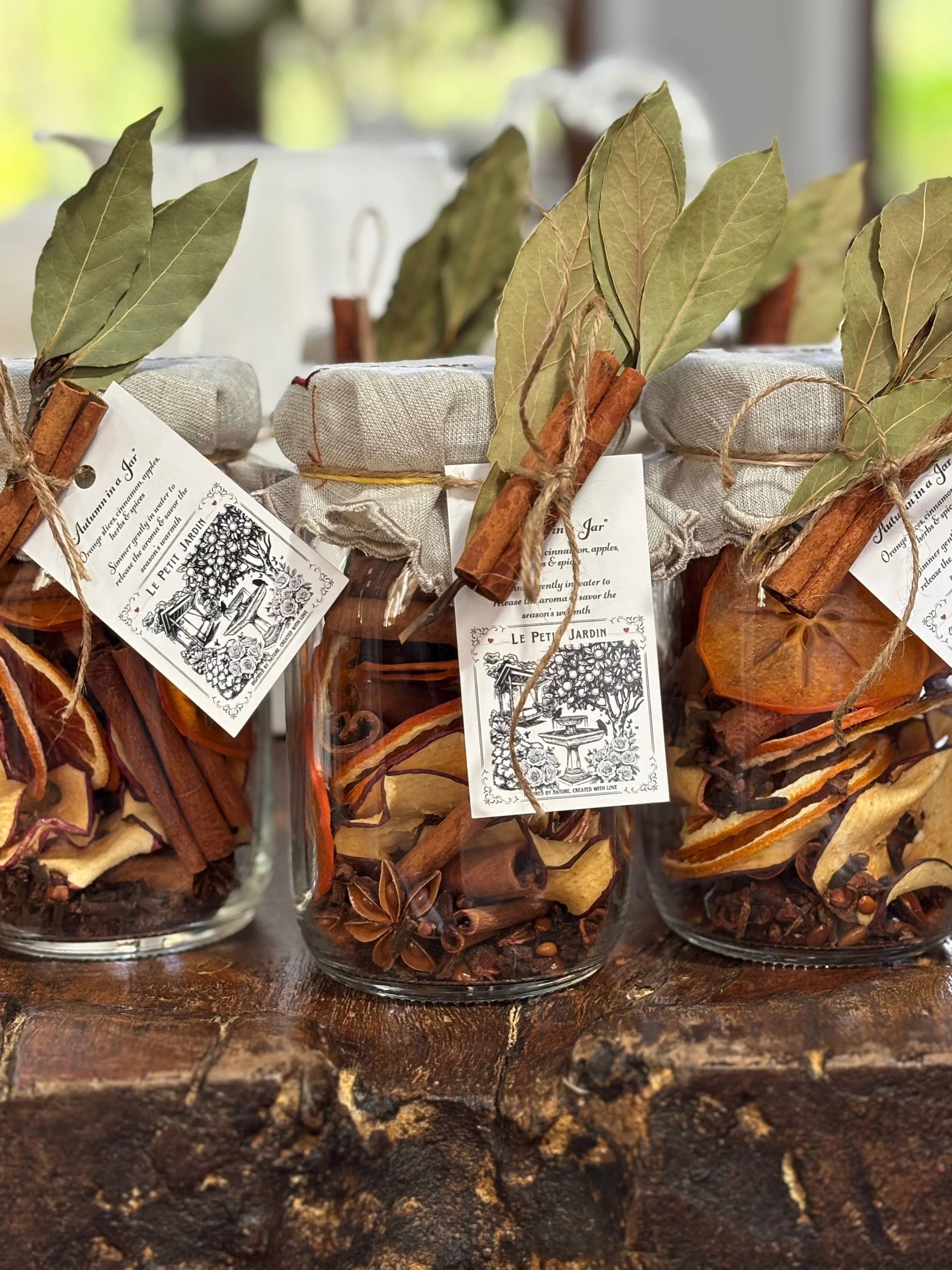 Three glass jars filled with dried fruit slices, cinnamon sticks, and star anise, tied with twine and decorated with dried leaves and cinnamon sticks, labeled with black and white vintage-style tags.
