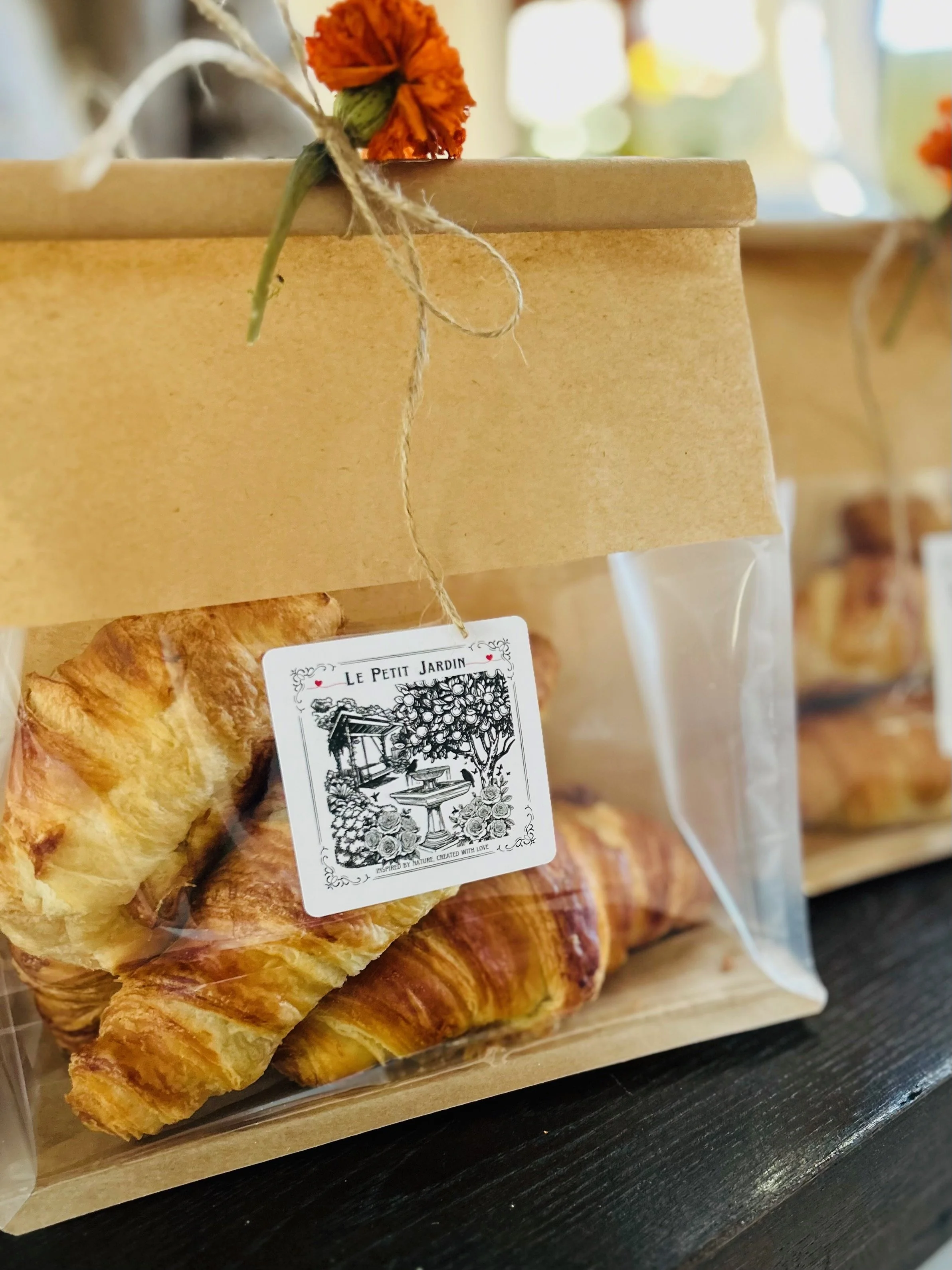 Baked croissants in a clear plastic bag with a brown paper label, decorated with an orange flower and tied with twine, sitting on a wooden surface.