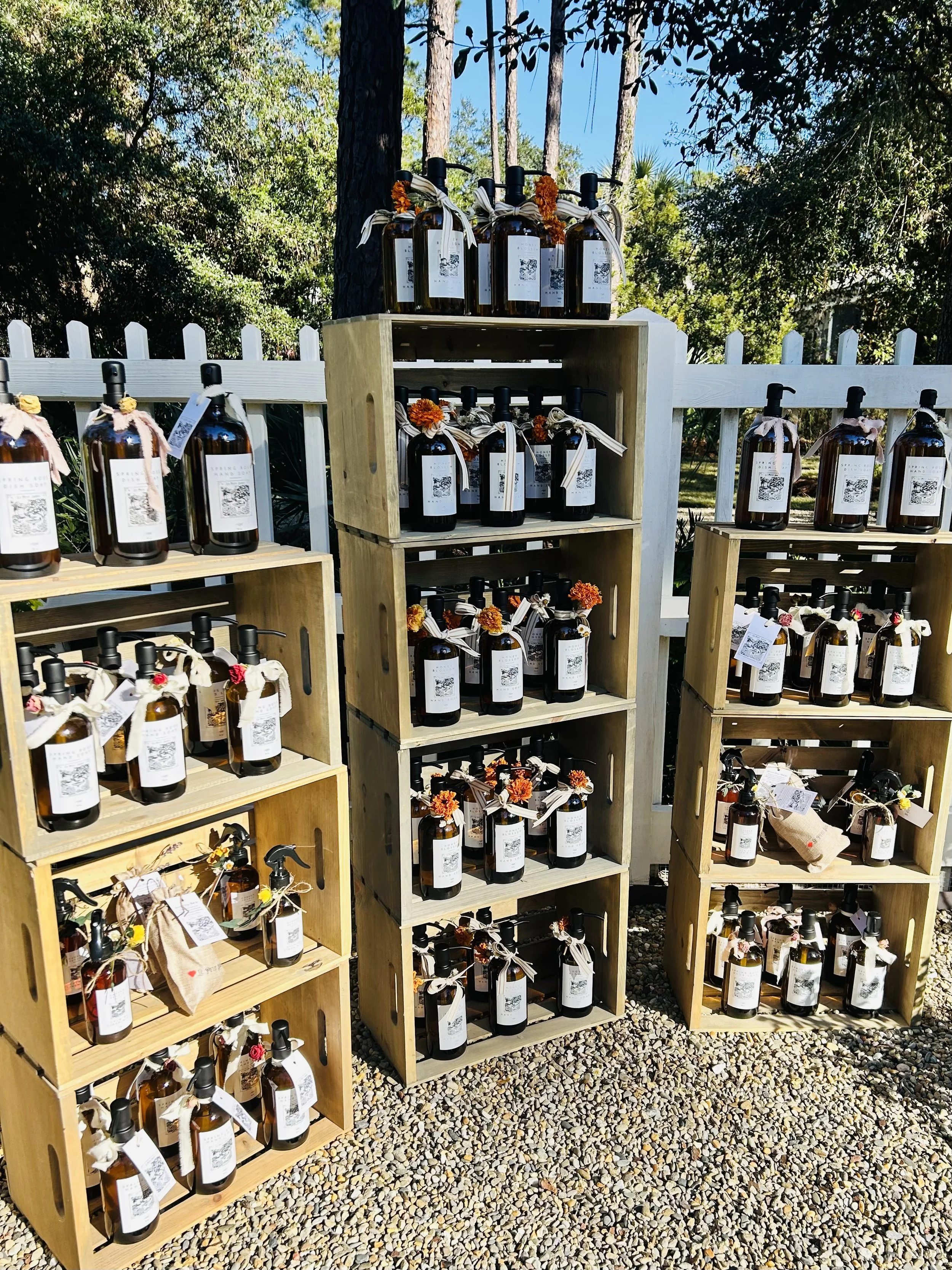 Display of amber glass bottles with pump dispensers and decorative ribbons and flowers, arranged on wooden crates outdoors with a white picket fence and trees in the background.