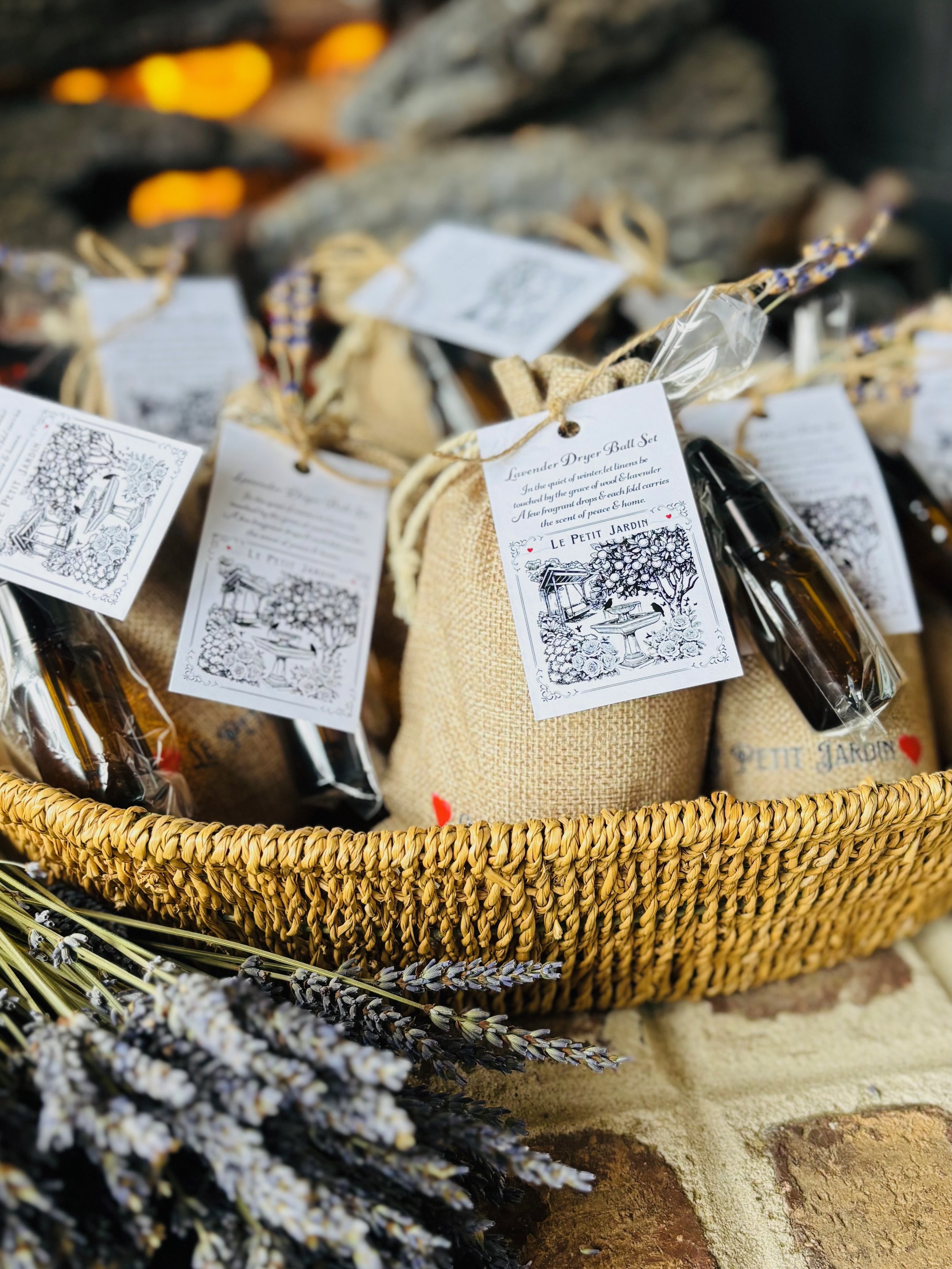 A woven basket filled with burlap sacks and small bottles, each with a tag labeled "Lavender Dryer Ball Set" and an illustration of a garden scene. Dried lavender stems are at the front in the foreground.