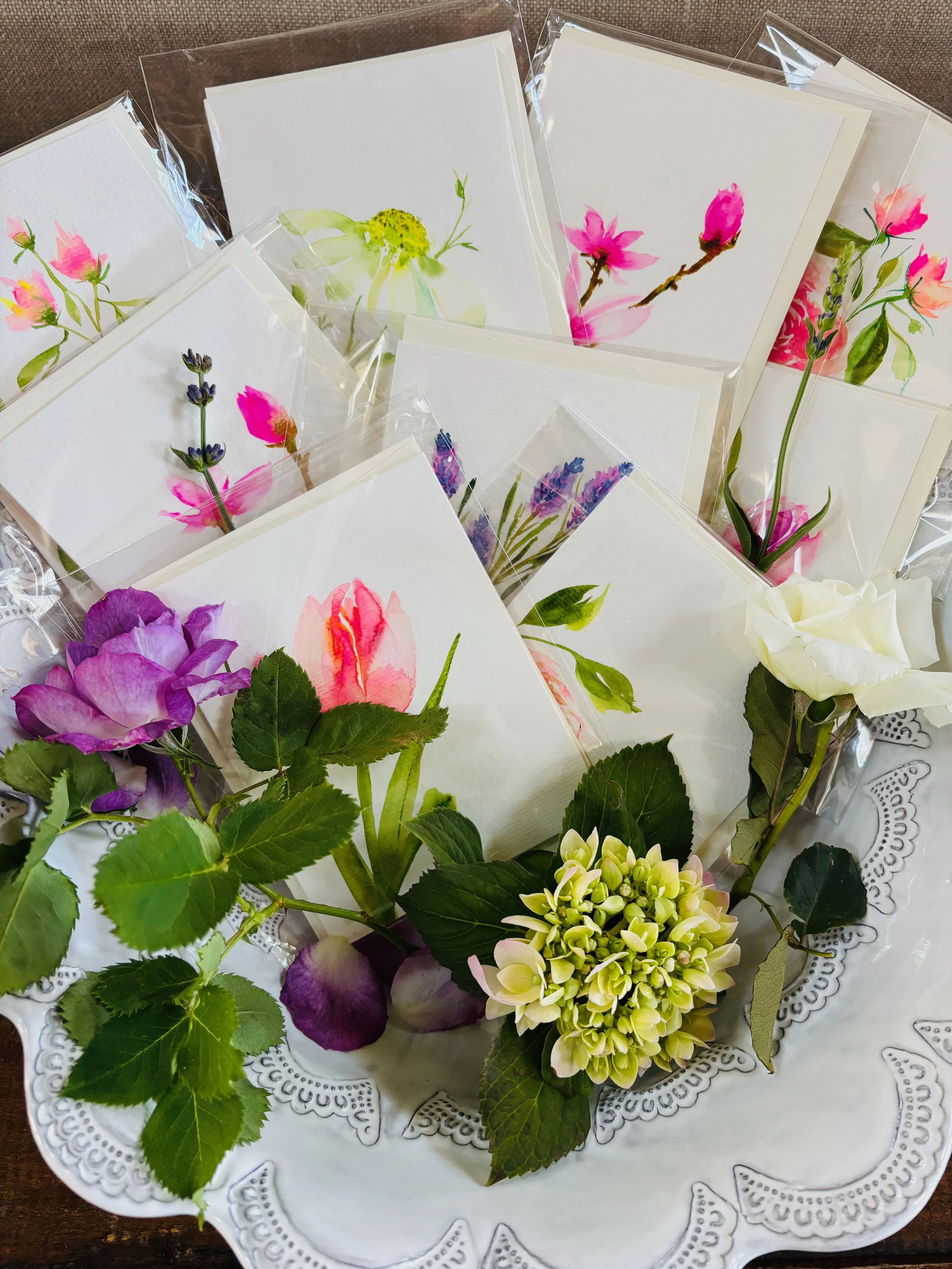 A decorative white ceramic tray holding assorted handmade watercolor floral greeting cards and fresh flowers, including purple, pink, and white blossoms, arranged for display.