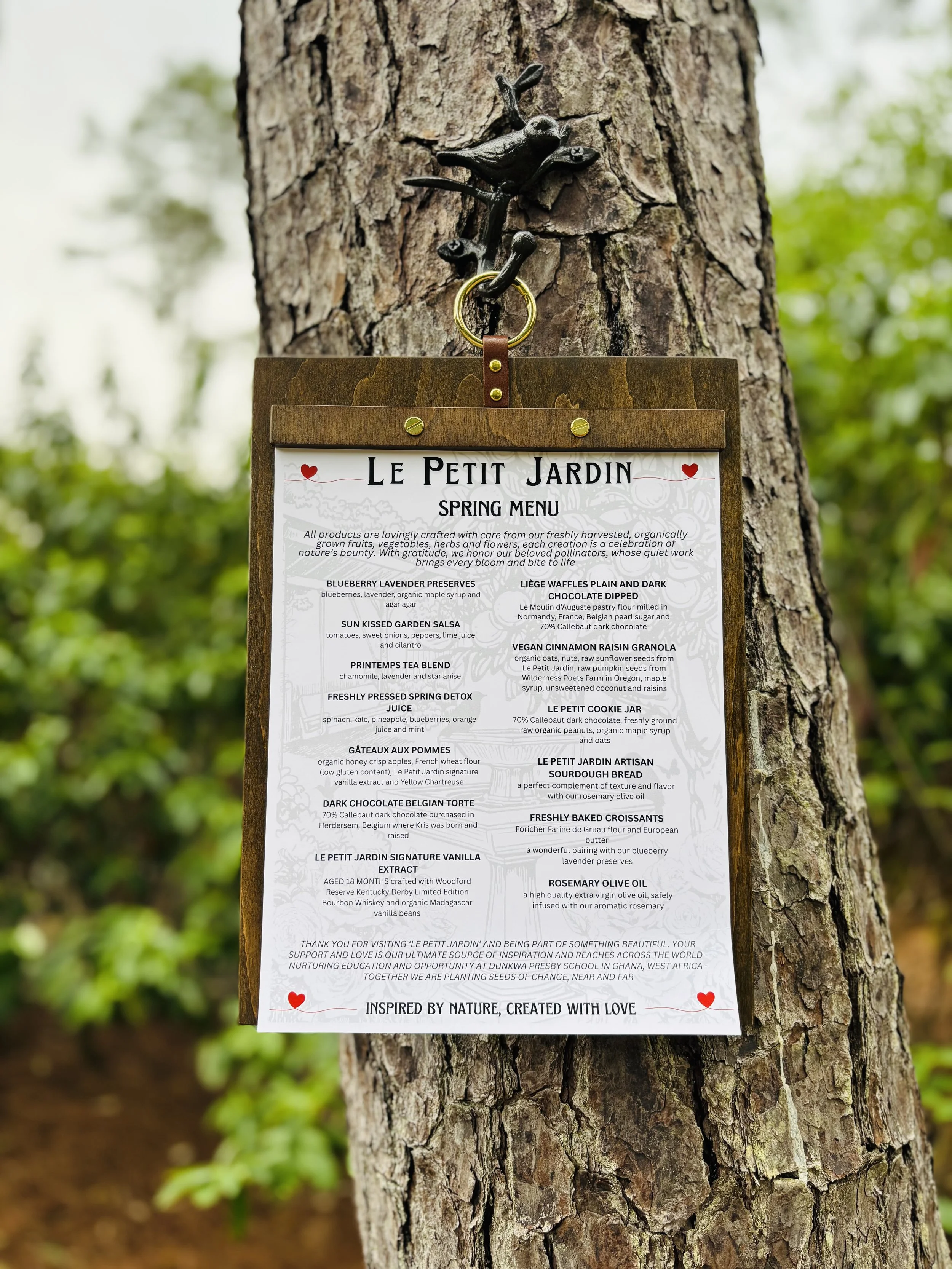 A printed menu titled Le Petit Jardin Spring Menu hanging on a tree trunk with a decorative hook in an outdoor setting.