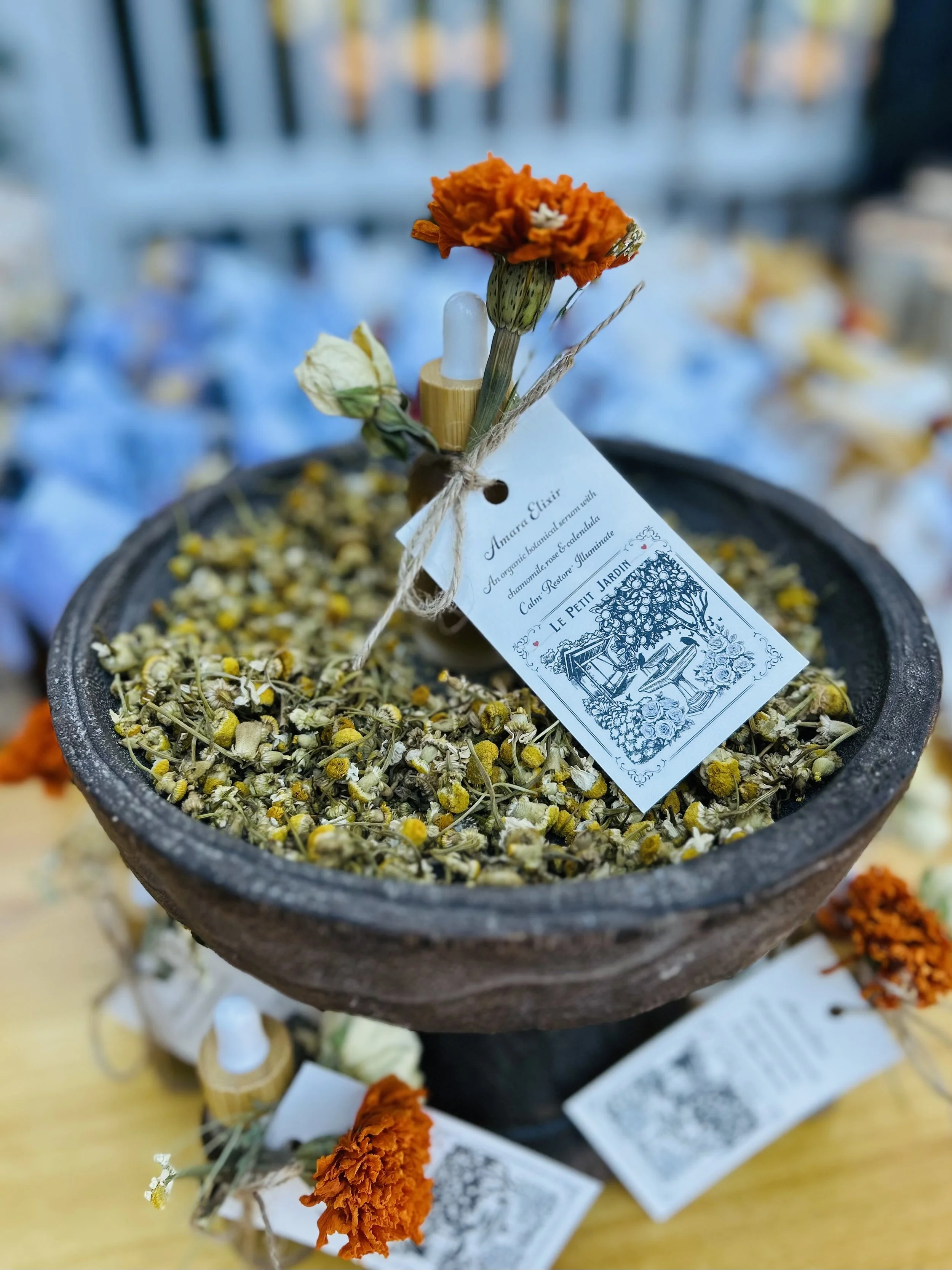 A round black bowl filled with dried chamomile flowers, with a small bouquet of orange, white, and green flowers attached to a dropper bottle and a tag labeled 'Amara Elixir' with a sketch of a garden scene. The background shows a fence and scattered