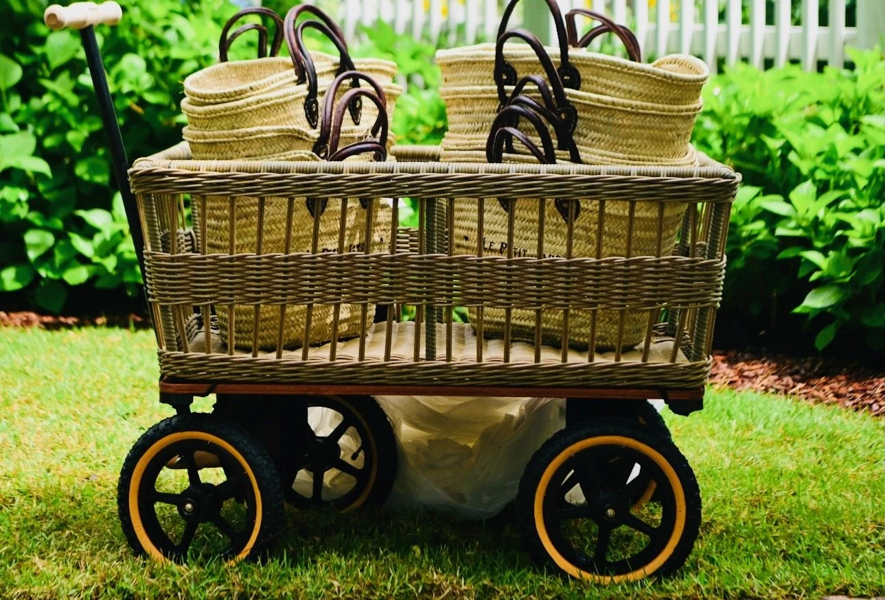 A wicker shopping cart with four woven handbags on a grassy lawn, with a white picket fence and green bushes in the background.