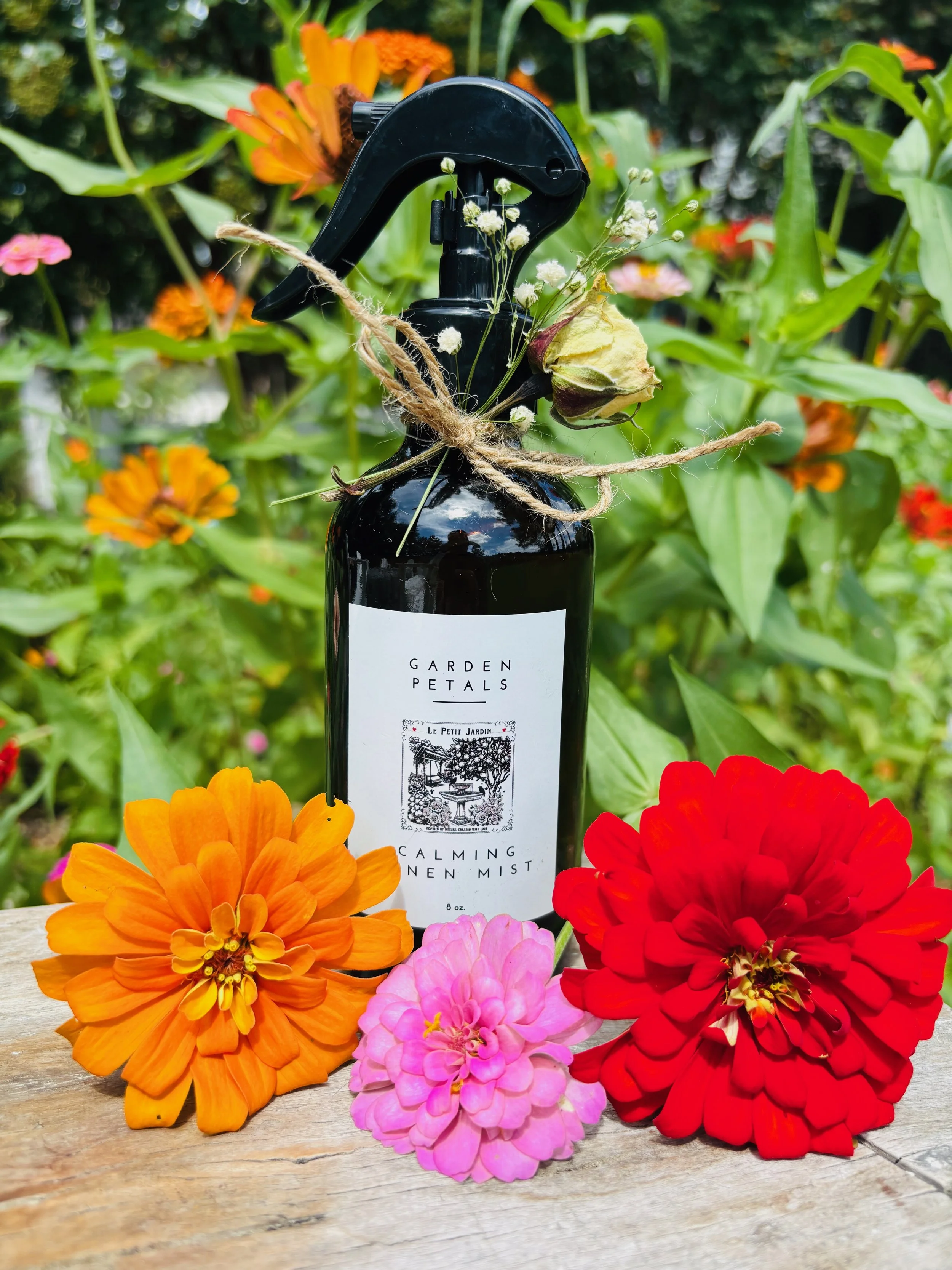 A black spray bottle labeled "Garden Petals Calming Linen Mist" is surrounded by colorful flowers, including orange, pink, and red zinnias, in a garden setting.