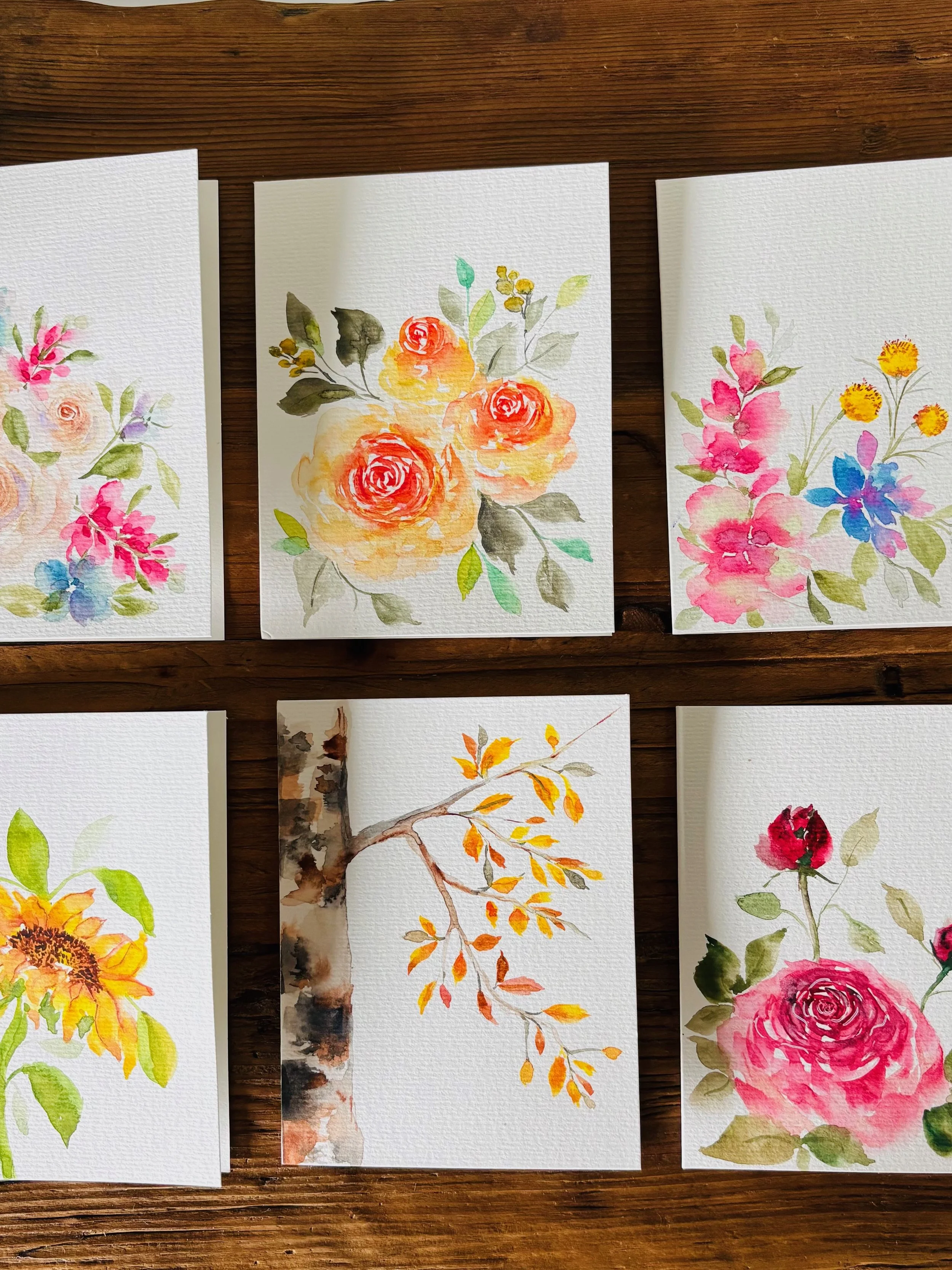 Six watercolor floral and nature paintings displayed on a wooden surface, including roses, sunflowers, a branch with orange leaves, and various pink, yellow, and colorful blooms.