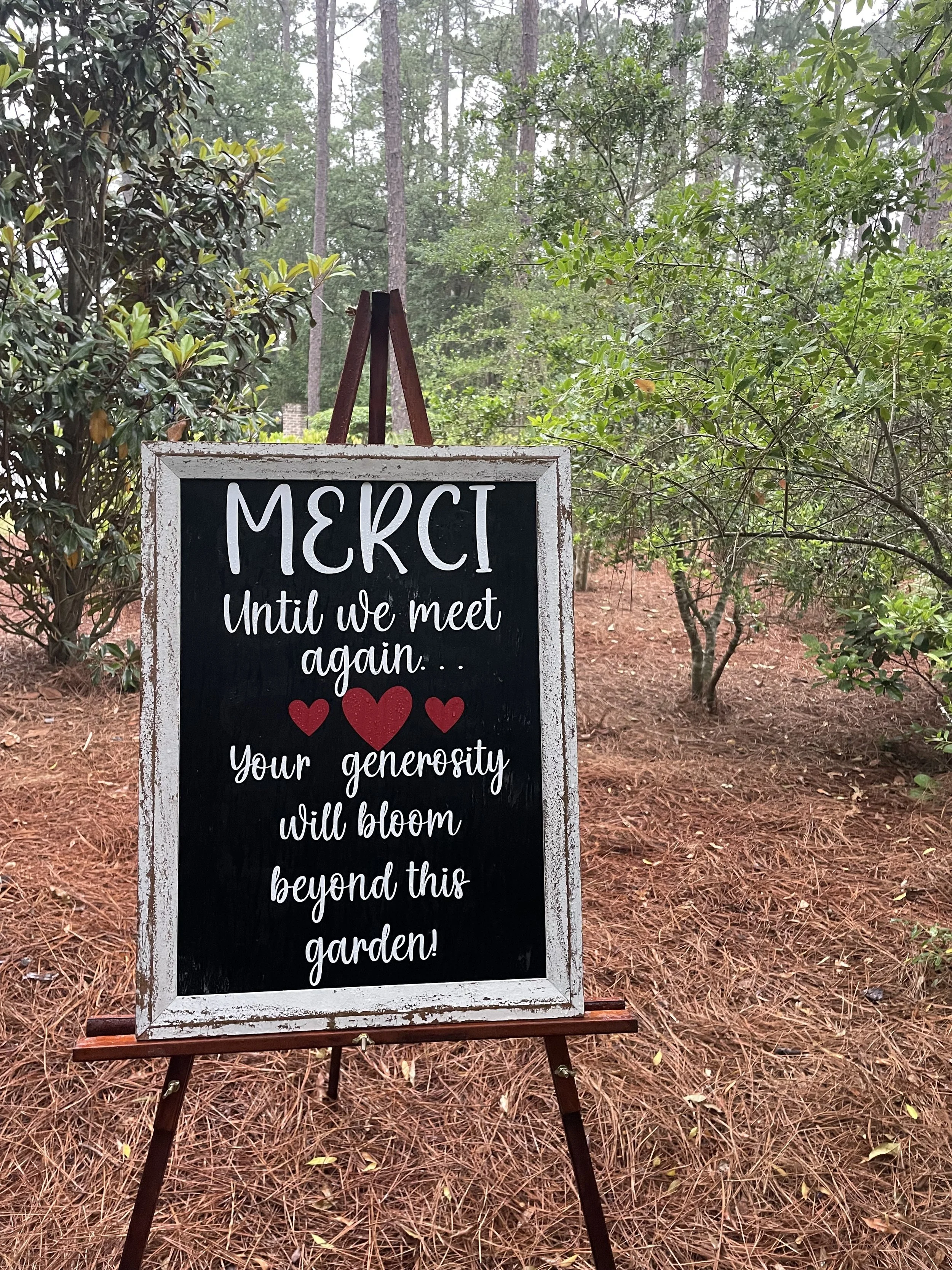 A chalkboard sign with a white frame on a wooden easel in a wooded garden, displaying a message of thanks with red hearts.