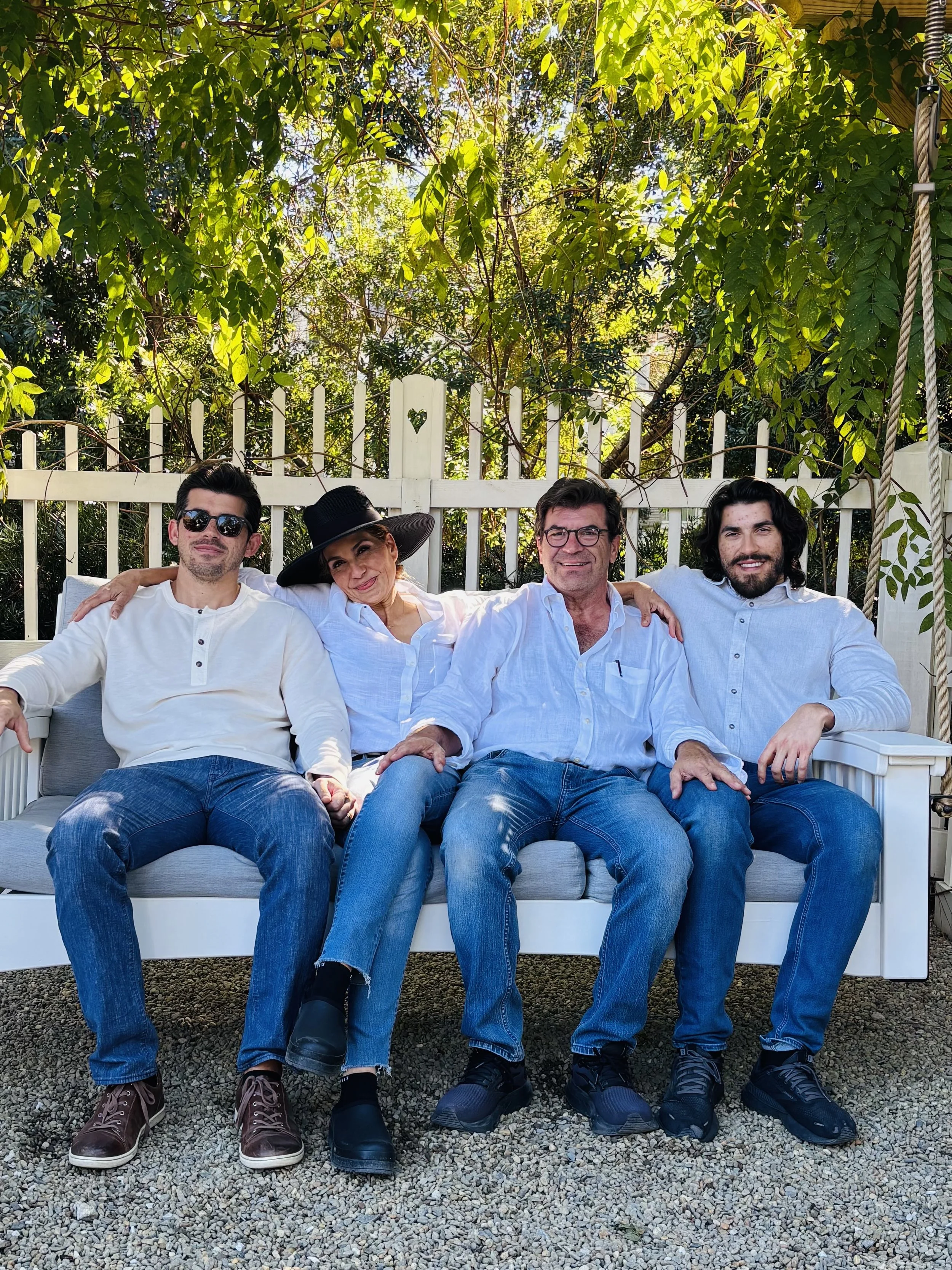 Four people sitting on a white outdoor bench in a garden, dressed casually, smiling, with greenery and a white fence in the background.