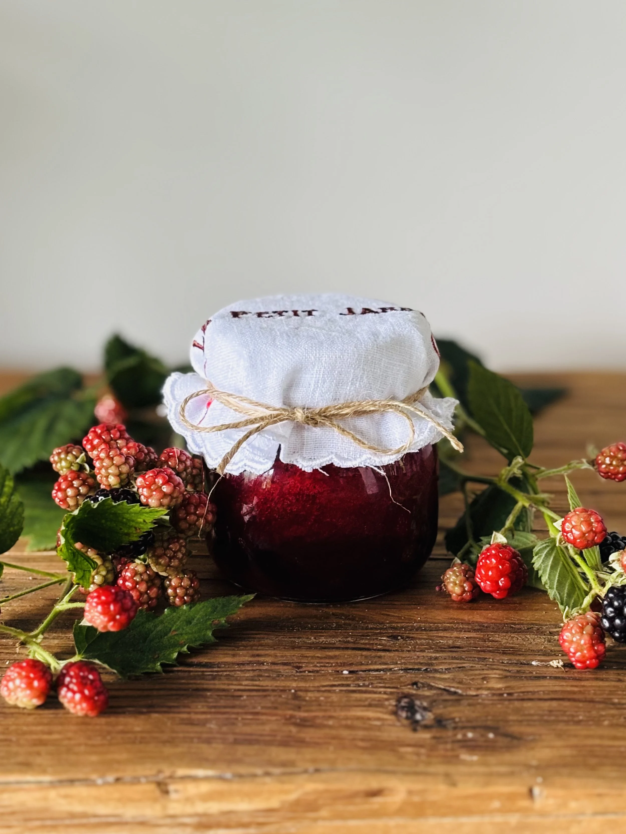 A jar of homemade blackberry jam with a white cloth cover tied with a string, surrounded by fresh blackberries and green leaves on a wooden surface.