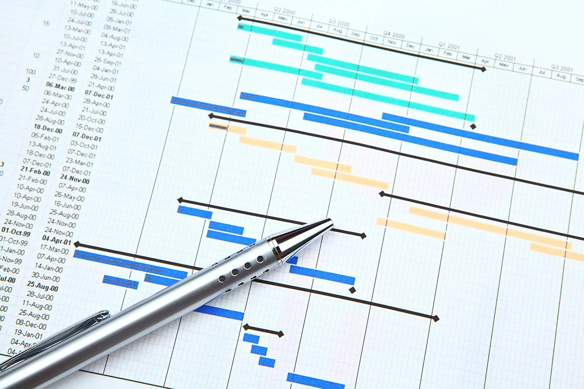 A printed project timeline or Gantt chart showing parallel tasks with different start and end dates, overlaid with a silver ballpoint pen
