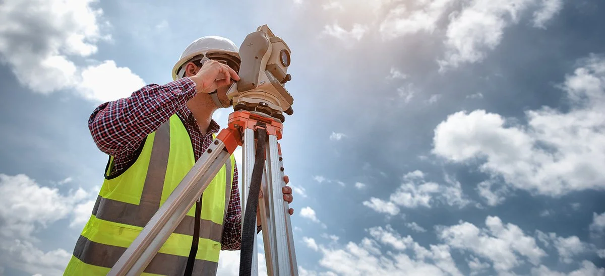 Surveyor in a yellow safety vest and white helmet using a theodolite on a tripod outdoors under a partly cloudy sky.