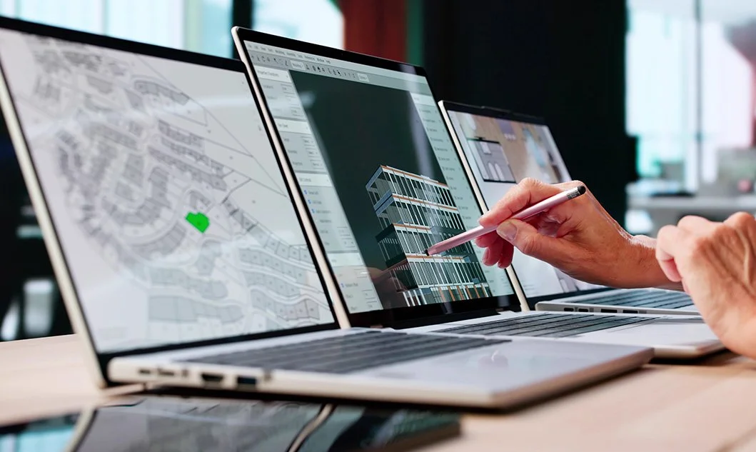 Three laptops displaying architectural designs and plans, with a person using a stylus to customize a building model on one screen.