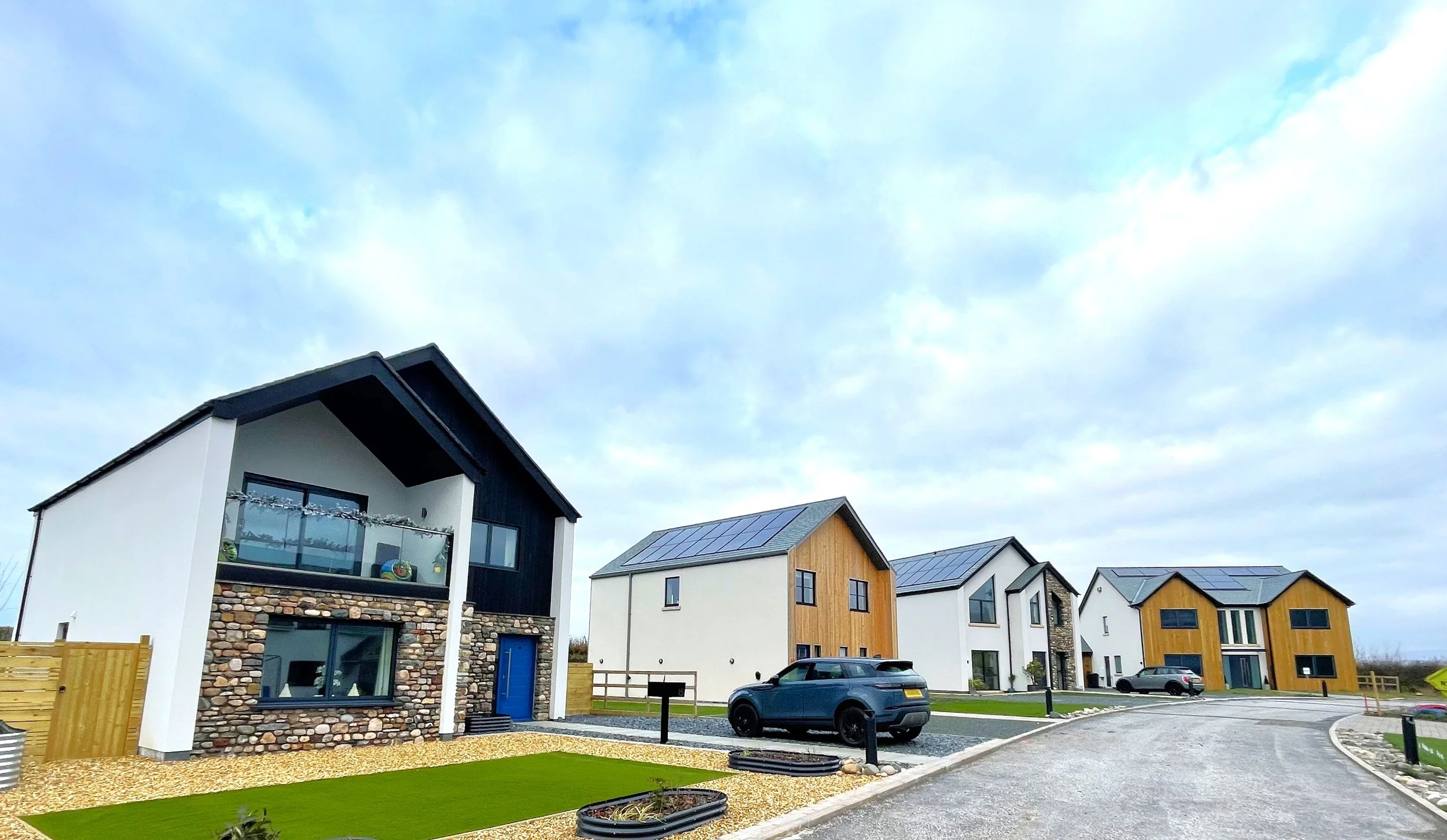 A row of modern suburban houses with solar panels on the roofs, parked cars, and a curved driveway on a cloudy day.