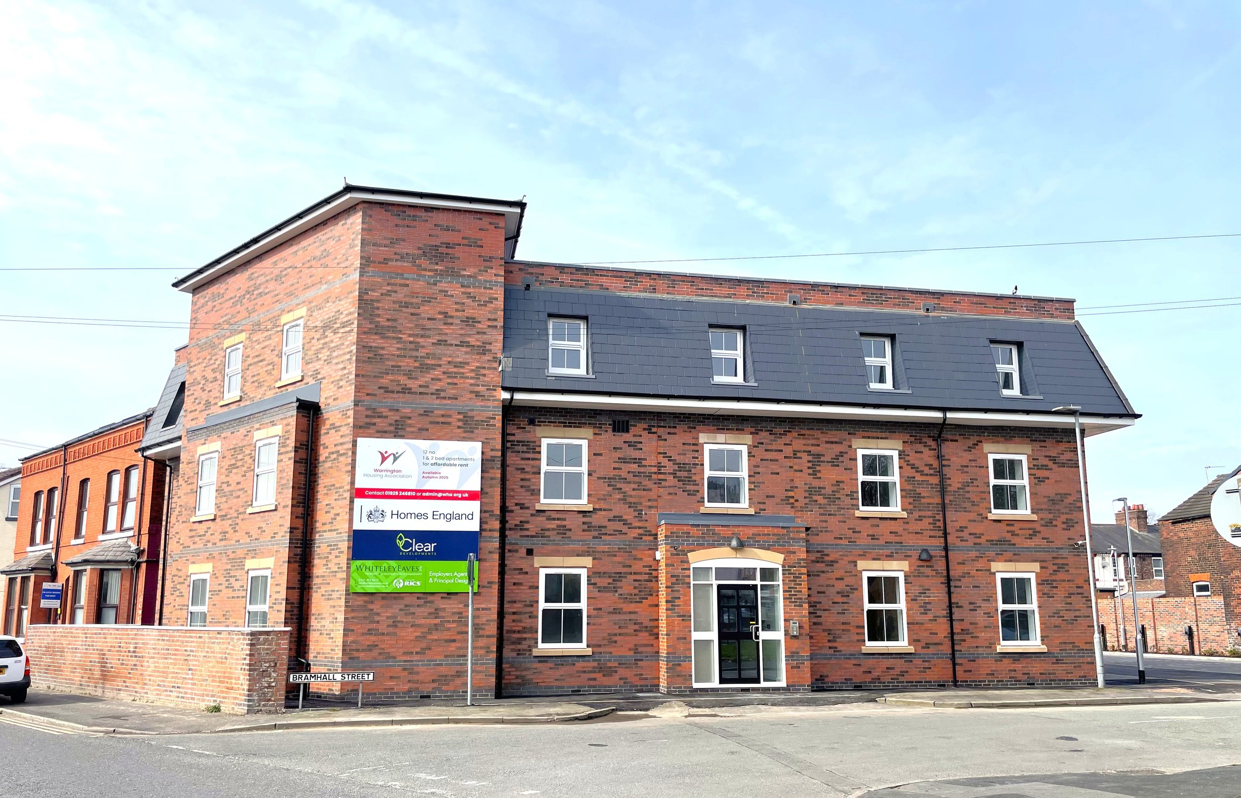 A three-story brick building on a street corner with multiple windows and a glass entrance door. There are signs on the side of the building indicating it's associated with housing and development companies. The street sign reads 'Bramhall Street,' a