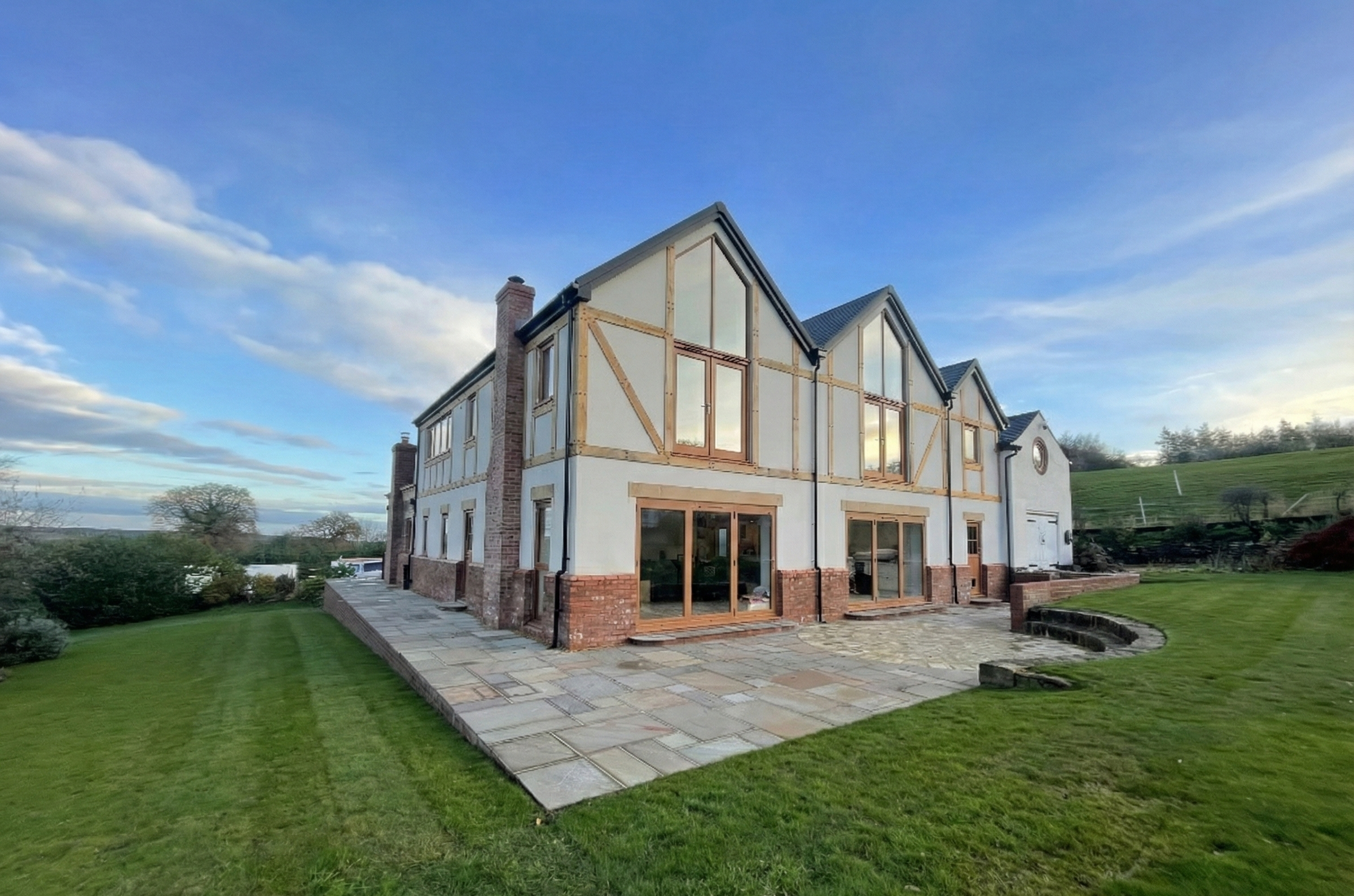 A two-story house under renovation with visible wooden framing on the upper level, brick accents, glass sliding doors and large windows, set in a landscaped yard with green grass and a stone patio, against a background of blue sky with clouds and a h
