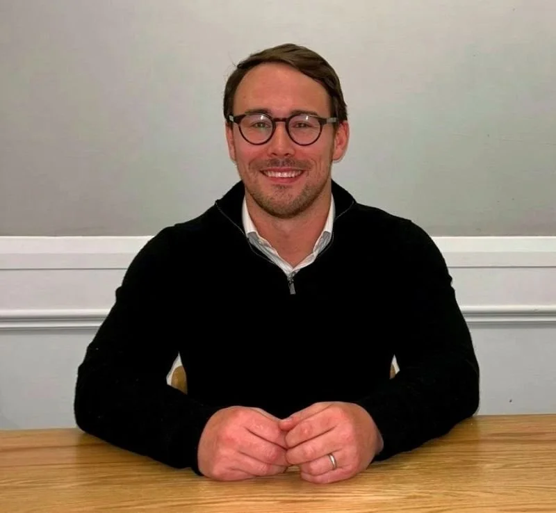 A man with glasses, beard, and short hair wearing a black zip-up sweater and a white collared shirt sitting at a wooden table, smiling, with a plain light-colored wall in the background.