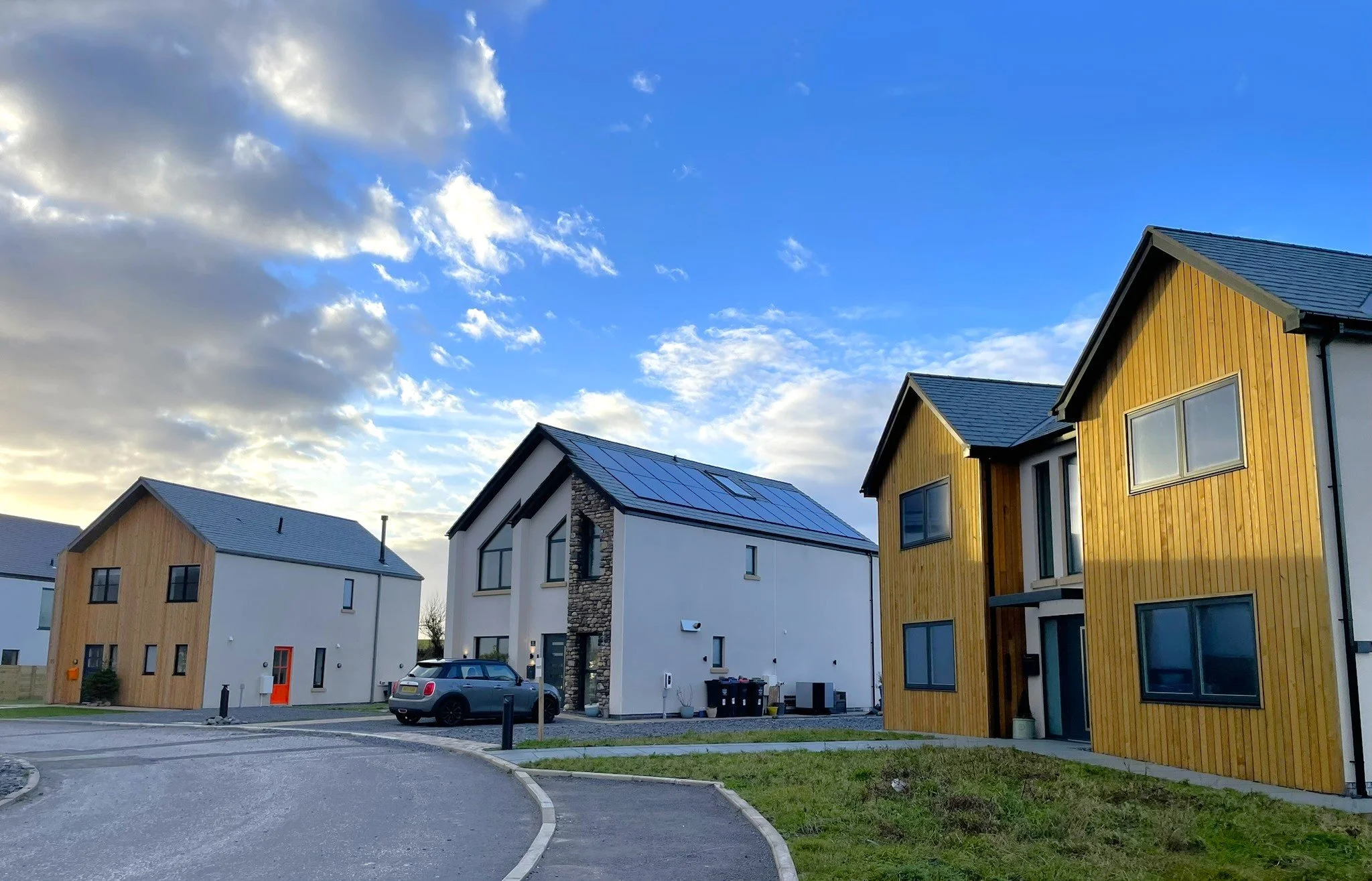 Modern residential houses with wooden and white exteriors, a driveway, cars, and a partly cloudy sky.